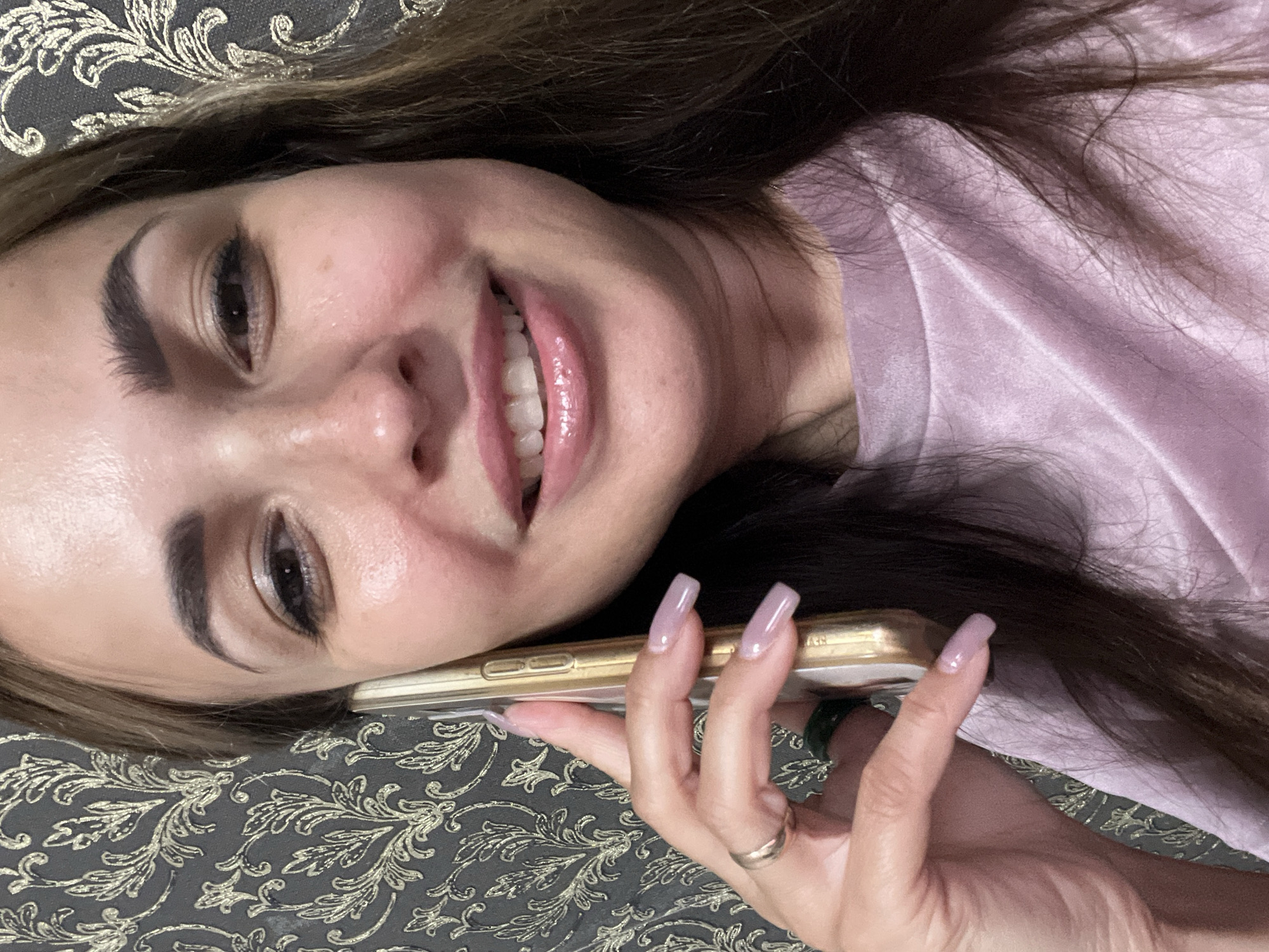 A woman with brown hair and light skin is shown in a close-up, selfie-style shot. She is holding a gold smartphone to her right ear with her left hand, which has long, light purple nails and two rings on her ring finger. She is wearing a light purple, velvety top. Her lips are slightly parted, revealing white teeth, and she is smiling. Her eyes are open, and she is looking towards the camera. The background consists of a dark, patterned wallpaper with a gold floral design. The lighting appears to be from a single source, creating some highlights on her skin and lips.