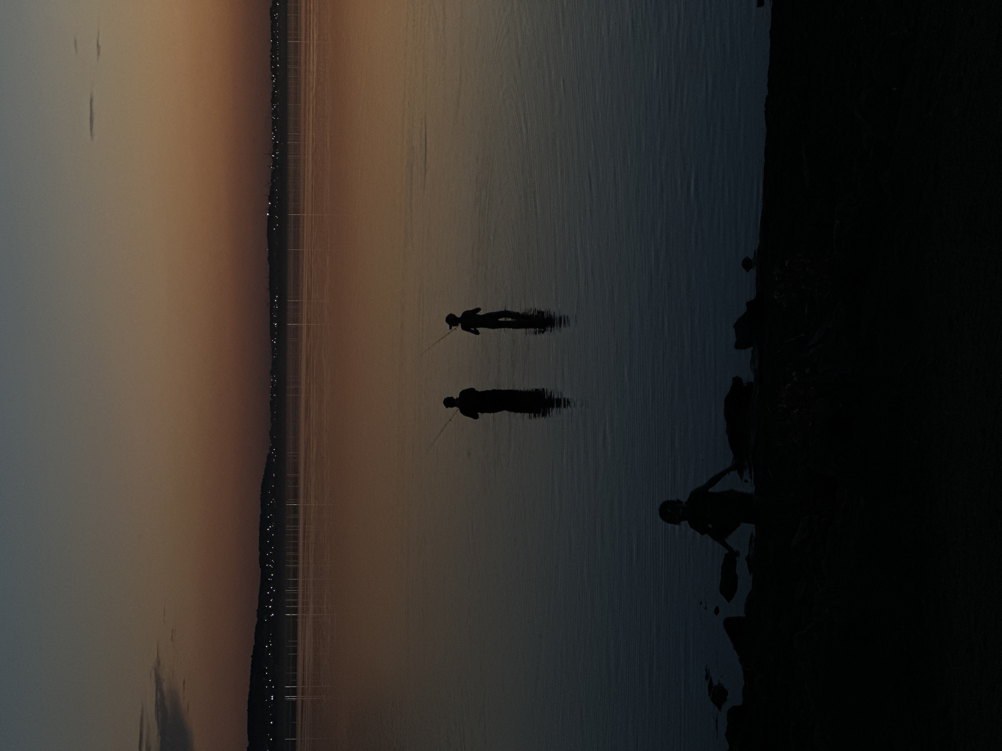 The image depicts a serene twilight scene over a body of water, likely located in the city of Kokshetau, Kazakhstan, given its geographical context. The sky displays a gradient of deep blues and warm oranges, characteristic of dusk, with a faint line of distant lights visible along a silhouetted treeline on the horizon. The water's surface is calm, reflecting the muted colors of the sky. In the middle ground, two silhouetted figures stand in the water, each holding a fishing rod, engaged in the activity of fishing. In the foreground, a third silhouetted figure, possibly a child, is visible near the rocky shoreline, appearing to be either playing or observing. The overall impression is one of peaceful solitude and quiet enjoyment of nature at the close of day.