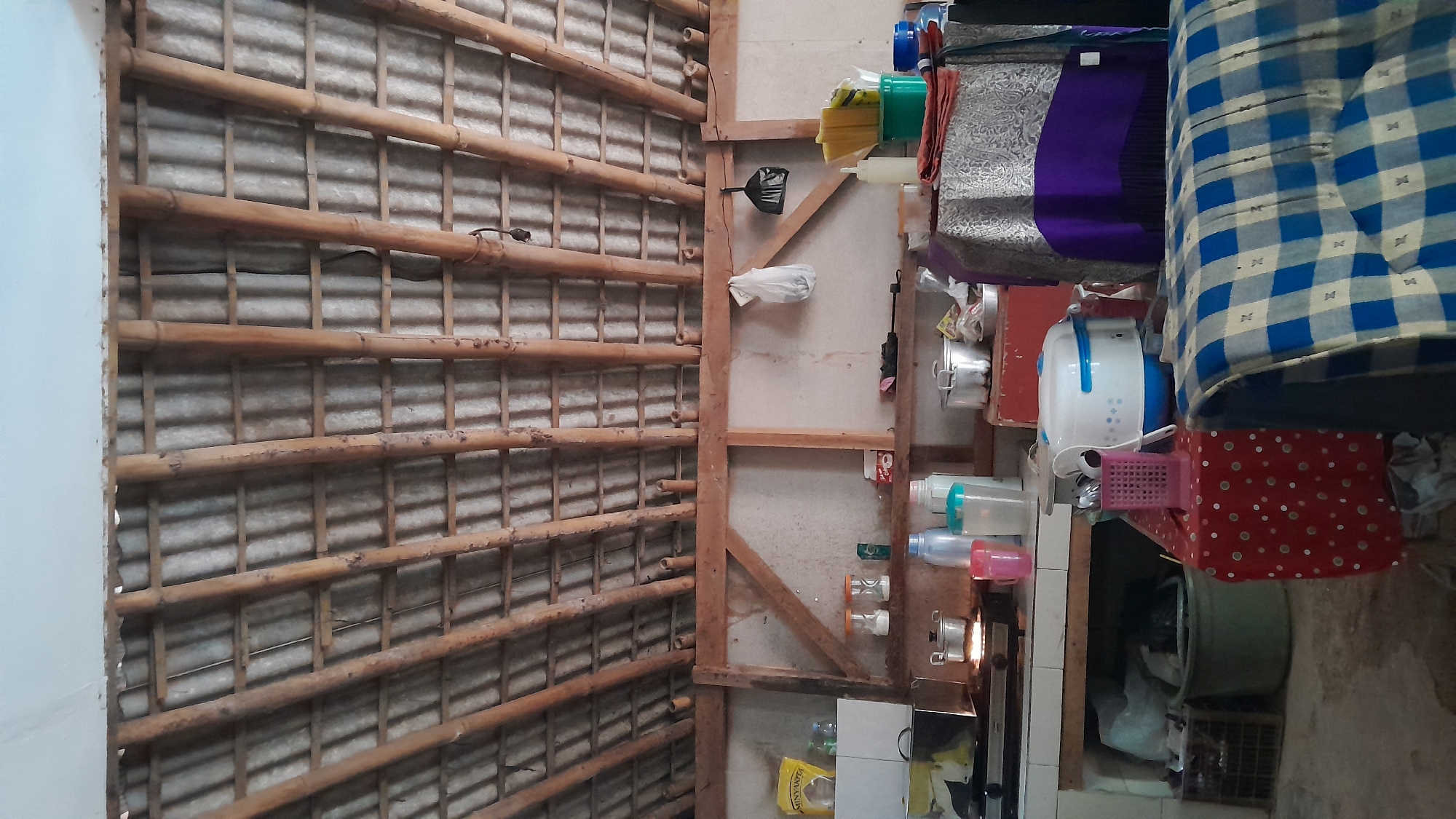 The image depicts the interior of a simple kitchen with a rustic, open-air feel, likely located in Sumedang, Indonesia. The ceiling is constructed from corrugated metal sheets supported by a framework of bamboo poles, a common building material in rural Indonesian homes. The walls appear to be plastered or made of a rough material.
In the foreground, a red tablecloth with white polka dots is laid out on a surface, topped with a white and blue rice cooker and a pink plastic utensil holder. To the right, a blue and white checkered fabric is draped over what appears to be a sofa or seating area.
The middle ground features a countertop with various kitchen items. A gas stove with pots on it is visible to the left. Several plastic bottles and containers, along with stainless steel pots and pans, are arranged on shelves and the countertop. A red box-like structure is also present.
In the background, shelves are stocked with jars, bottles, and other kitchen supplies. Two plastic bags, one white and one black, hang from a wooden beam. The overall impression is of a functional, well-used kitchen in a dwelling that prioritizes natural and readily available materials for construction.