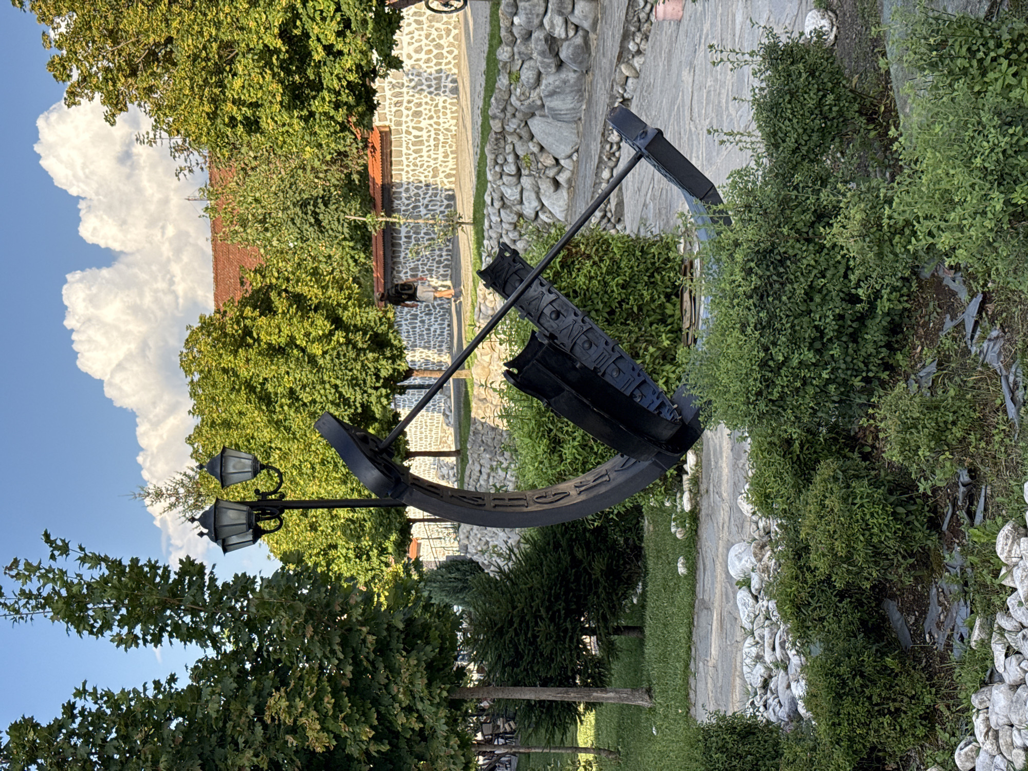In the city of Bansko, Bulgaria, a large, dark-colored anchor sculpture is prominently displayed in an outdoor setting. The anchor, made of metal, features intricate details and appears to be part of a monument or public art installation. It is situated in a landscaped area with green grass, various bushes, and a stone walkway. In the background, a person wearing a backpack and shorts is walking away from the camera. Behind them, a building with a red-tiled roof and a stone wall is visible. Two black lampposts stand to the left of the anchor sculpture. The sky above is blue with fluffy white clouds, and several trees with green foliage surround the scene. The overall environment suggests a public park or a well-maintained outdoor space.