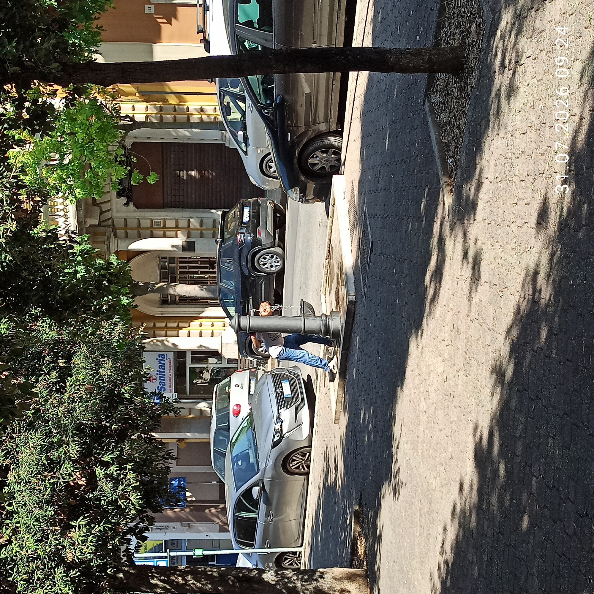 This is a medium-shot, eye-level photograph taken on a sunny day in Modugno, Italy, as indicated by the timestamp "31.07.2026 09:24" in the lower right corner. The scene captures a street with parked cars, a public water fountain, and a person interacting with it.
In the foreground, the pavement is made of interlocking grey bricks, partially covered by the dappled shadows of trees. A thin tree trunk is visible on the right side of the image.
In the middle ground, a dark grey public water fountain stands on a raised concrete base. A person, wearing a grey t-shirt and blue jeans, is bending over the fountain, appearing to drink or collect water. The person's left hand is near the spout. Water is actively flowing from the fountain's spout, creating a spray.
Several cars are parked on the street. On the left, a silver hatchback (likely a Peugeot 208) is parked. Further back, a dark-colored Jeep SUV is visible, and behind it, a lighter-colored hatchback. To the right, a dark-colored sedan is parked, partially obscured by the tree trunk.
In the background, a building with architectural details like arched doorways and columns is visible. A sign that reads "Sanitaria" with a red cross symbol is mounted on the building's facade, suggesting a healthcare-related establishment.
The overall impression is a slice of everyday life on a street in an Italian city.