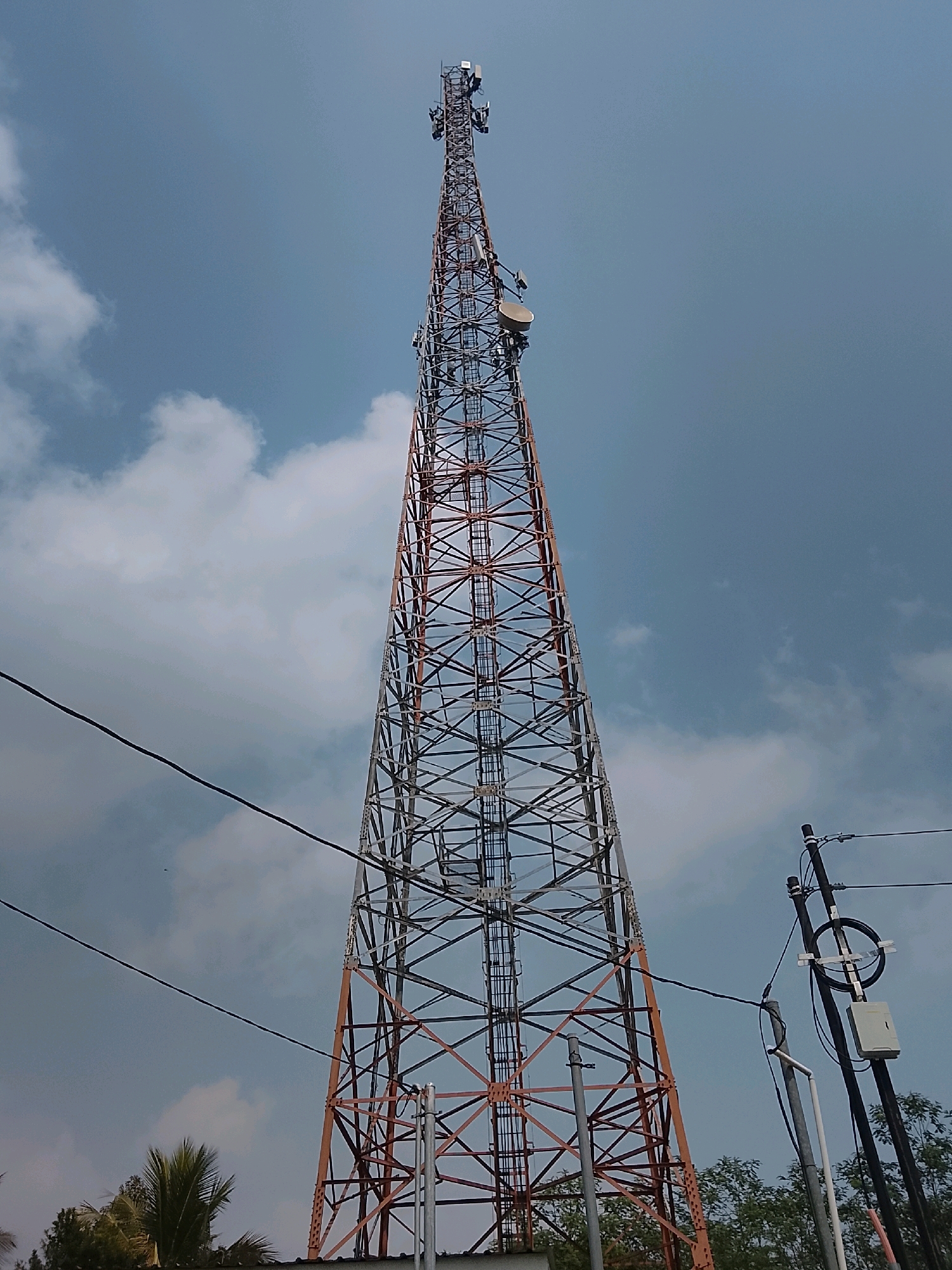 A tall, lattice cell tower, painted orange and silver, dominates the frame, extending upwards into a pale blue sky dotted with white clouds. Several antennas and dishes are affixed to the tower at various heights. In the foreground to the right, a smaller utility pole is visible, supporting electrical wires and a junction box. Power lines also stretch diagonally across the left side of the image. Below the tower and pole, glimpses of trees and foliage are visible, suggesting an outdoor, potentially semi-rural environment. The overall scene depicts infrastructure for telecommunications.