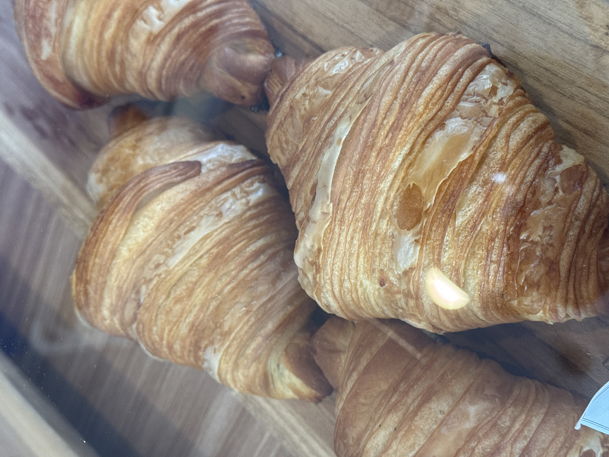 The image displays a close-up view of several golden-brown croissants resting on a light wooden surface. The croissants, exhibiting a classic crescent shape with visible flaky layers, appear freshly baked and have a glossy, caramelized sheen in some areas. One prominent croissant occupies the right side of the frame, showcasing its intricate texture and rich color. Other croissants are visible behind and to the left, arranged in a casual cluster.
The scene is viewed through a clear barrier, likely glass or plastic, which creates reflections and slightly diffuses the sharpness of the image. A distinct, bright, elongated reflection of light is visible on the surface of the largest croissant, hinting at a light source in the environment. The wooden surface below the pastries is a warm, light brown, suggesting a natural material like a display shelf or tray.
The image captures a display of baked goods, characteristic of a bakery or cafe setting in North Kuta, Indonesia. There is no activity taking place other than the static display of the food. No people are visible. A small, white, unreadable piece of paper or tag is partially visible in the bottom right corner, obscured by a croissant and the barrier. The overall impression is one of delicious, fresh pastries.