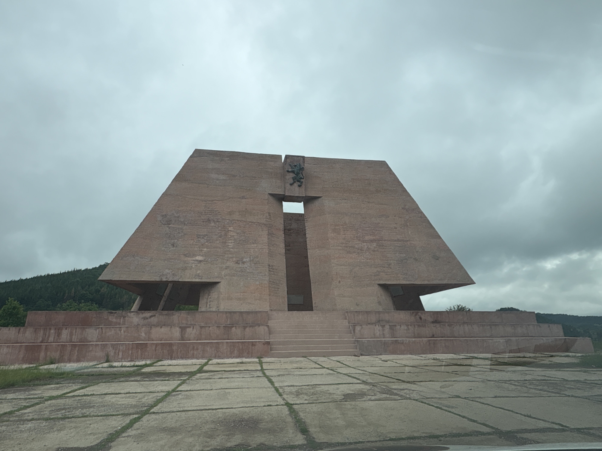 The image shows the Buzludzha Monument in Gurgulyat, Bulgaria. The monument is a Brutalist architectural structure made of concrete and brick, with a distinctive pyramid-like shape. It is situated on a hilltop, surrounded by lush green trees and rolling hills. The sky is overcast, with heavy clouds creating a dramatic atmosphere. In the foreground, a paved area with a grid pattern of concrete slabs and grass patches leads up to the monument. The monument itself is the focal point, with its imposing scale and stark design. A bronze lion sculpture is mounted on the facade of the monument, above a central opening. The monument appears weathered and shows signs of neglect, consistent with its abandoned status.