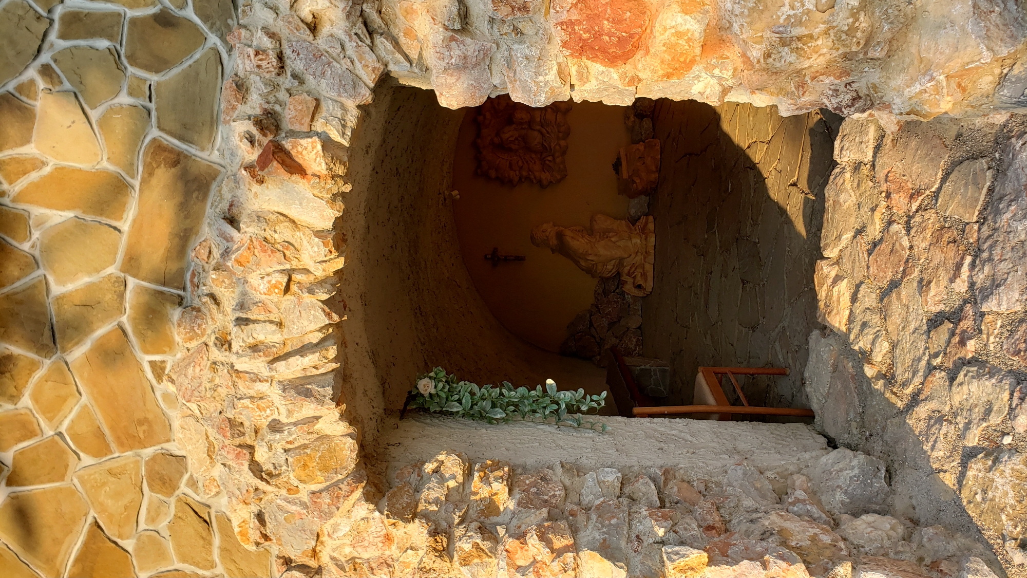 The image depicts an arched stone entrance leading into a small, dimly lit grotto. The exterior is constructed from rough-hewn, irregular stones in shades of beige, orange, and brown, with visible mortar. Sunlight casts strong shadows on the ground in front of the entrance, illuminating a path of similarly irregularly shaped stone paving.
Inside the grotto, the walls and ceiling are also stone, forming a curved arch. A small, dark wooden chair is visible to the left, beside a hanging artificial green vine with a white rose. Further inside, a statue of a kneeling figure, likely religious in nature, is placed on the stone floor. Above the statue, a crucifix is mounted on the wall. To the right of the statue, a carved relief is affixed to the wall. The floor of the grotto is paved with flat, irregular stones. The lighting suggests either late afternoon or early morning sun.
The location is identified as the St. Nicholas Cathedral (Konkatedrála sv. Mikuláša) in Prešov, Slovakia, specifically its outdoor Stations of the Cross.