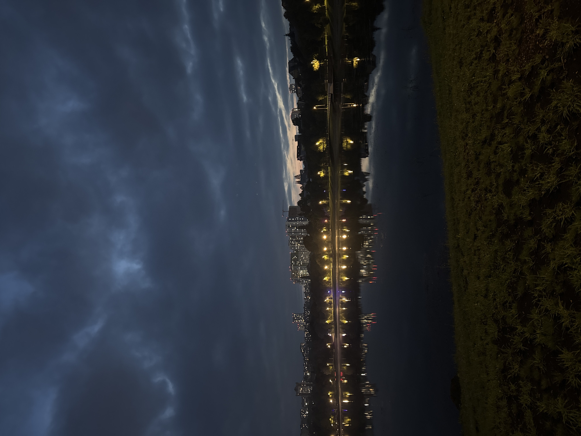 The image depicts a nighttime scene along the bank of a body of water, likely a lake or reservoir, in Ufa, Russia. The sky is dark and cloudy, with some faint streaks of light suggesting the end of twilight or early dawn. In the background, a city skyline is visible, illuminated by numerous building lights. The lights are reflected on the surface of the water, creating a shimmering effect. The foreground consists of a grassy slope leading down to the water's edge. The scene appears peaceful and tranquil.