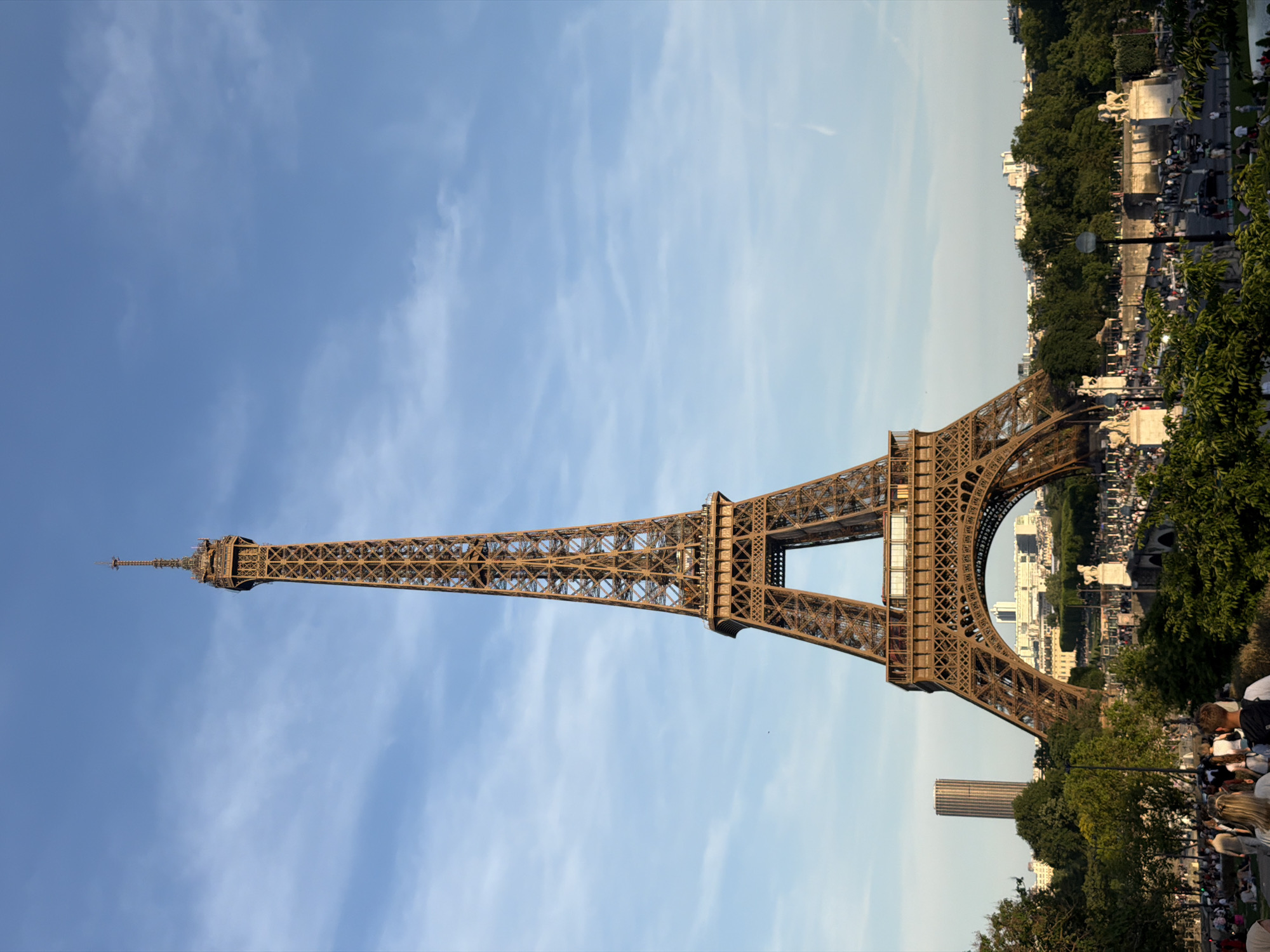The Eiffel Tower, a wrought-iron lattice tower on the Champ de Mars in Paris, France, dominates the frame, towering above the cityscape. The tower's intricate ironwork is visible against the blue sky with streaks of clouds. At its base, crowds of people gather, likely tourists. Lush trees and greenery add to the scene's visual texture. In the distance, other buildings and monuments of Paris are visible.