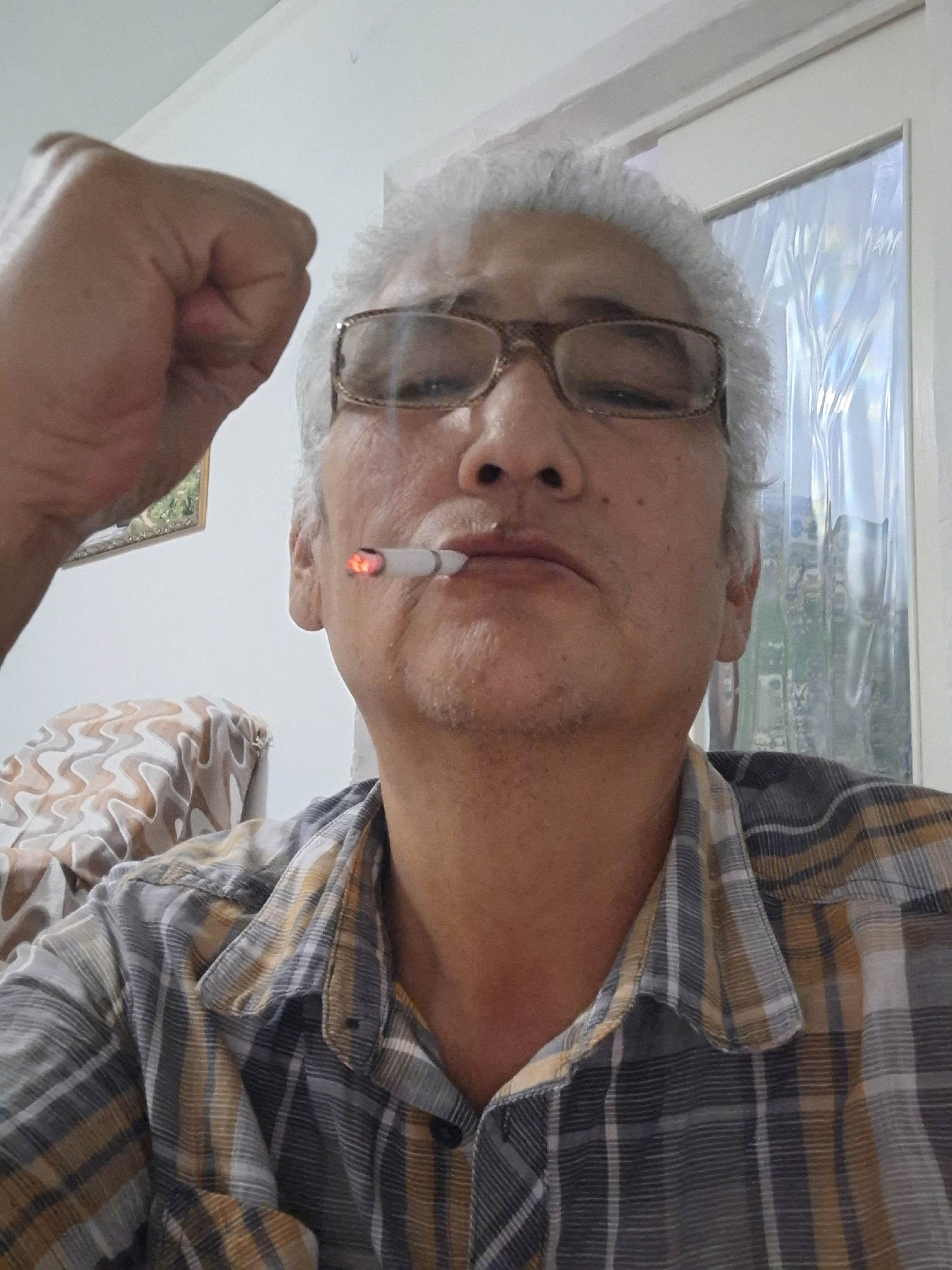 An elderly man with gray hair and glasses is sitting indoors, smoking a cigarette. His left fist is raised in the air. He is wearing a plaid shirt. In the background, there is a window with a patterned curtain and a framed picture on the wall. The scene appears to be in a residential setting, likely an apartment, given the interior elements. There is no specific indication of the location within the city of Almaty, Kazakhstan.