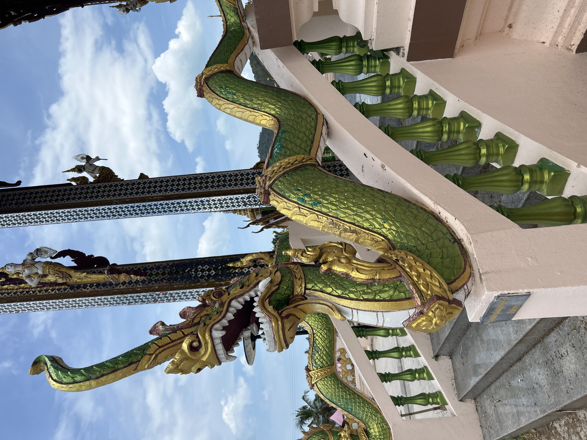 This image depicts an outdoor scene, likely a temple or shrine, in the Ko Samui District of Thailand. The foreground is dominated by a green and gold serpent statue, an ornate balustrade feature, that wraps around a set of stairs. The statue has a detailed head with an open mouth revealing sharp teeth, and its body is scaled and coiled. The balustrade posts are a vibrant green, supporting a pale pink railing.
In the background, the sky is a bright blue with scattered white clouds. Tall, decorative pillars with mosaic patterns and small figures adorn the sides of the structure. Palm trees and a distant hillside are visible, suggesting a tropical environment. The stairs themselves appear to be made of concrete and have a slightly worn, wet appearance, indicating recent rain or cleaning. A small, blue sign is affixed to the side of the railing near the bottom of the stairs. The overall impression is one of a culturally rich and visually striking location.