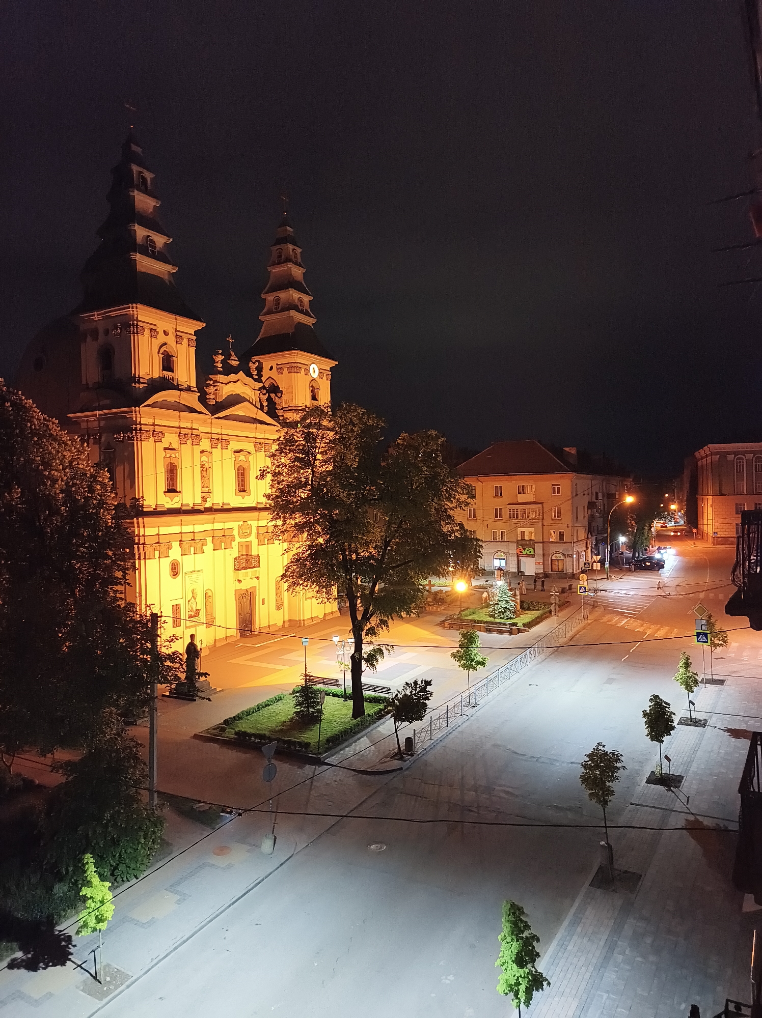 The image shows an elevated nighttime view of a section of Ternopil, Ukraine. A large, illuminated church with two prominent towers is the main focal point on the left side of the frame. The church is brightly lit, highlighting its architectural details. In front of the church is a square with a statue and landscaped areas. A road intersection is visible in the center of the frame, with crosswalks and traffic signs. Streetlights illuminate the road, revealing the pavement and sidewalks. There are trees planted along the sidewalk. In the background, other buildings can be seen, including multi-story structures. A few vehicles are visible on the road in the distance. The sky is dark, suggesting it is nighttime. No people are easily discernible.