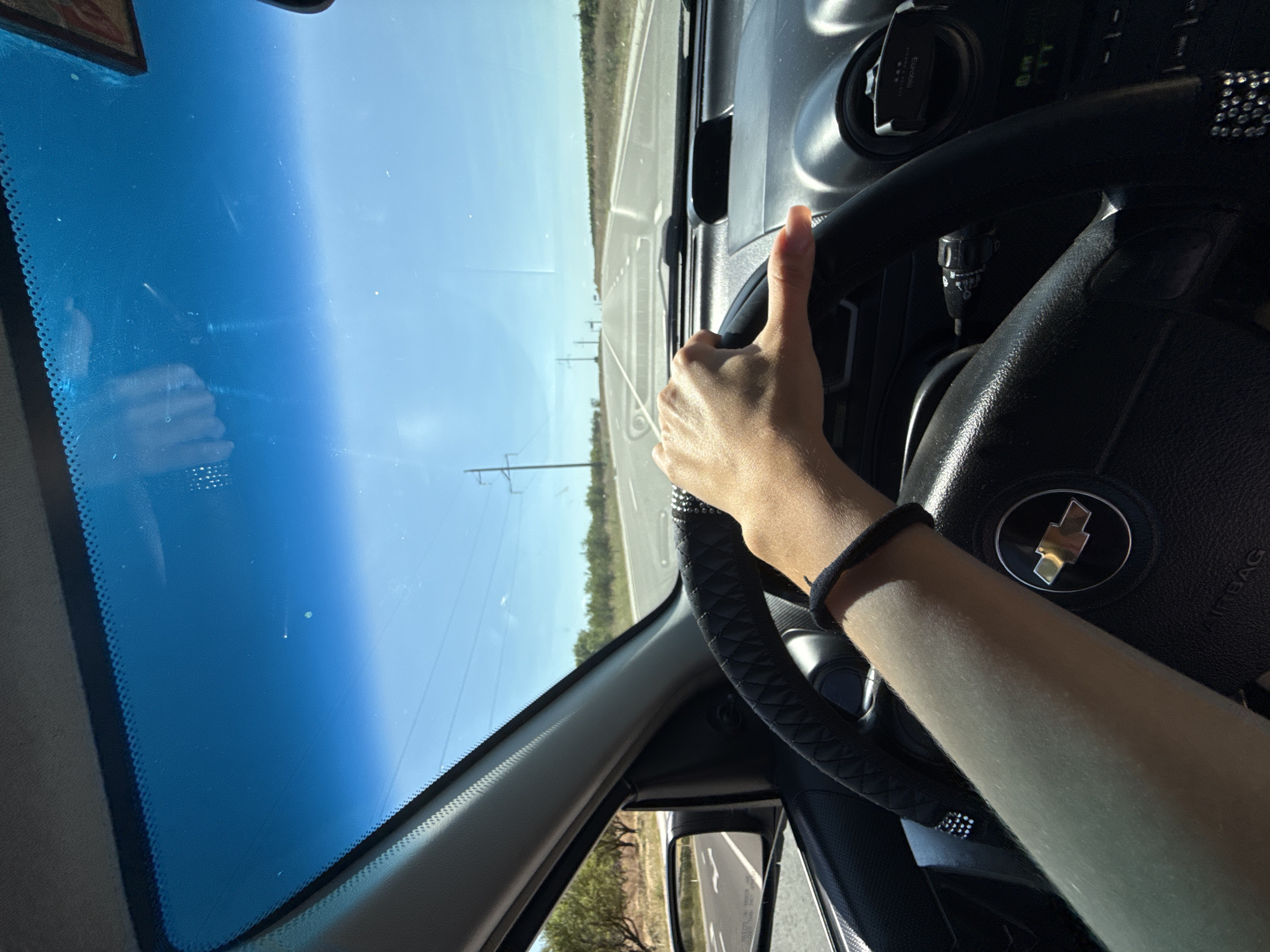 The image shows a first-person perspective of someone driving a car on a sunny day. The driver's hand is visible, resting on the steering wheel, which has a black cover with a diamond-like trim. The dashboard of the car is also visible, including a Chevrolet logo on the steering wheel and a car mount attached to the air vent. The road ahead is straight, with power lines and trees lining the sides. The sky is clear and blue.
The provided location of Cioc-Maidan, Moldova, is an agricultural village in the autonomous region of Gagauzia. Based on the visual cues in the image, such as the flat terrain, agricultural fields, and power lines, it is plausible that the scene is set in a rural or semi-rural area, consistent with the characteristics of Cioc-Maidan and its surroundings. However, the image itself does not contain any specific landmarks or features that definitively identify it as being within Cioc-Maidan itself.