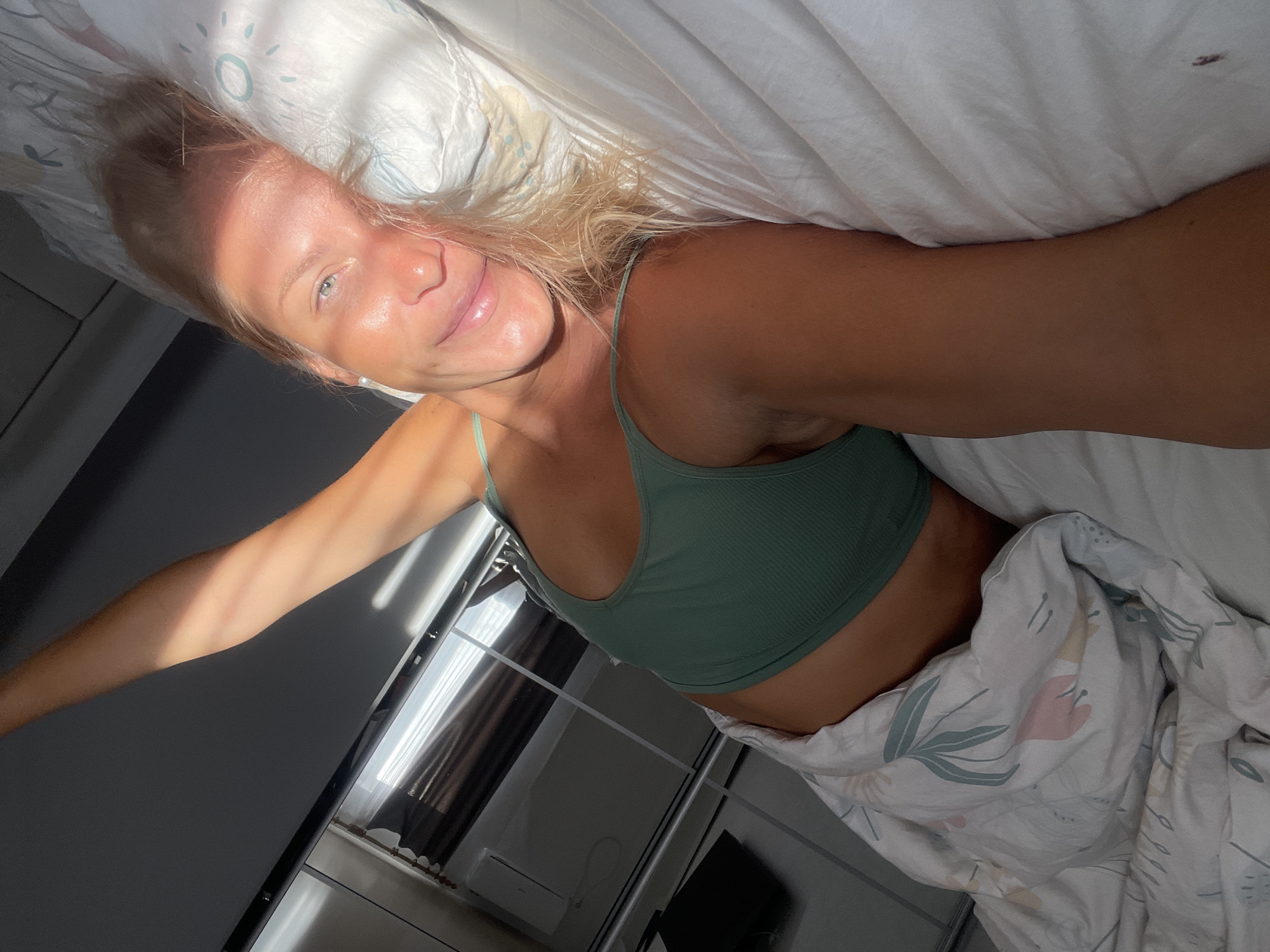 A blonde woman lies in bed, propped up on her elbow, with her right arm extended upwards. She is wearing a green tank top. Sunlight streams in from the right side of the image, casting shadows and highlighting her face and arm. The bed is covered in white sheets and a duvet with a floral pattern. The background shows a window with curtains and a sliver of an air conditioning unit. The scene appears to be in a bedroom in Baranovka, Russia.