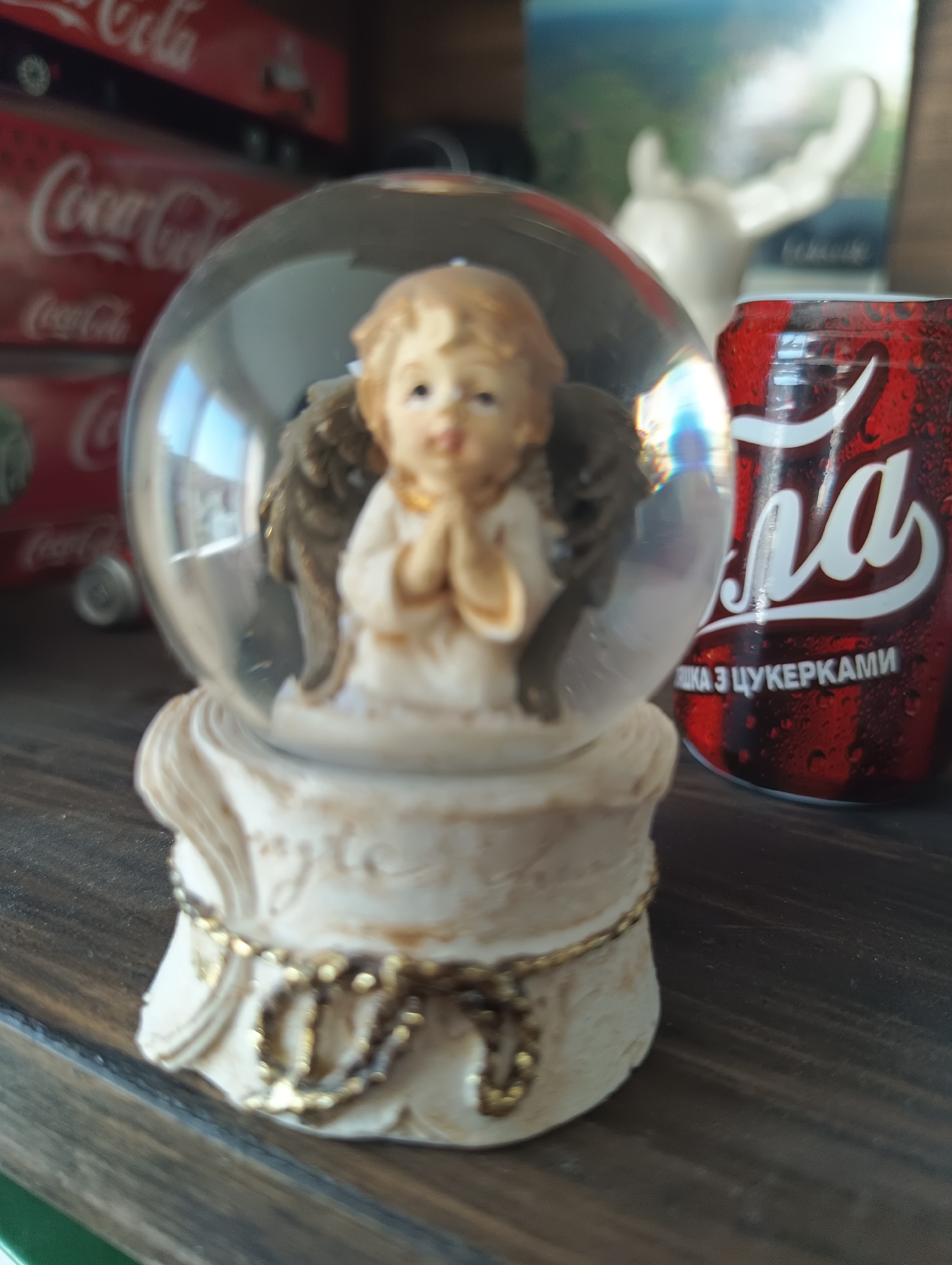 This is a close-up, eye-level, indoor photograph of a snow globe and a can of Coca-Cola. The snow globe, positioned in the foreground and slightly to the left, contains a small figurine of a praying angel with golden wings. The angel is dressed in a white robe and has light blonde hair. The base of the snow globe is intricately designed with a creamy, off-white finish and adorned with a delicate golden ribbon or cord.
To the right of the snow globe is a red can of Coca-Cola, partially visible. The can has water droplets on its surface, suggesting it is cold. Visible text on the can in white lettering reads "Coca-Cola" and below it, "НА ЗЦУКЕРКАМИ".
In the background, several stacked red Coca-Cola boxes are visible, with the "Coca-Cola" logo clearly discernible on them. Further in the background, a blurry white sculpture resembling an outstretched hand or antlers can be seen against a backdrop that appears to be an image or poster with a landscape.
The setting appears to be a shelf or tabletop, likely in a home or a shop. The lighting is soft, suggesting an indoor environment, possibly during the day. The overall mood is calm and perhaps slightly nostalgic, with the snow globe and familiar branding evoking a sense of childhood or simple pleasures. There are no clear indicators of time of day, weather, or specific emotions beyond the contemplative pose of the angel. The location context is Karnobat, Bulgaria, which is suggested by the text on the Coca-Cola can, likely in Bulgarian.