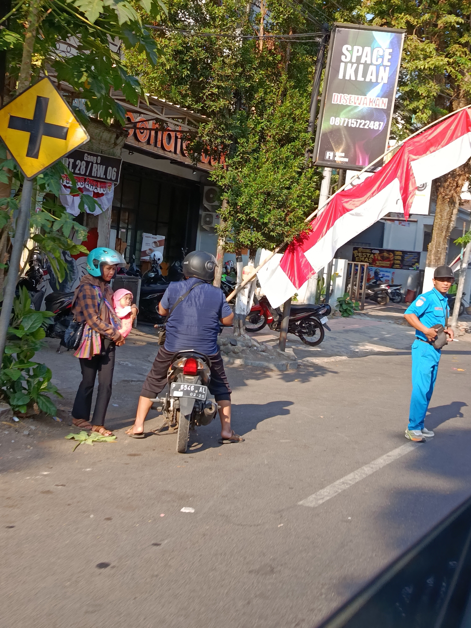 An outdoor daytime street scene in Bondowoso, Indonesia. A traffic sign indicating a crossroads is visible on the left side of the frame, accompanied by a smaller sign reading "GANG 0 RT. 28 / RW. 06". A banner displaying the Indonesian flag is draped across poles. In the foreground, a woman wearing a headscarf and a checkered shirt is standing, holding a baby wrapped in a pink blanket. A man on a motorcycle, wearing a dark blue shirt and black pants, is seated on the motorcycle, facing away from the camera. To the right, a person in a blue uniform, likely a traffic officer or security guard, is standing and holding a dark object. Several motorcycles are parked in the background, and trees and a building with an awning are also present. The street appears to be paved, with a white line marking a lane.
