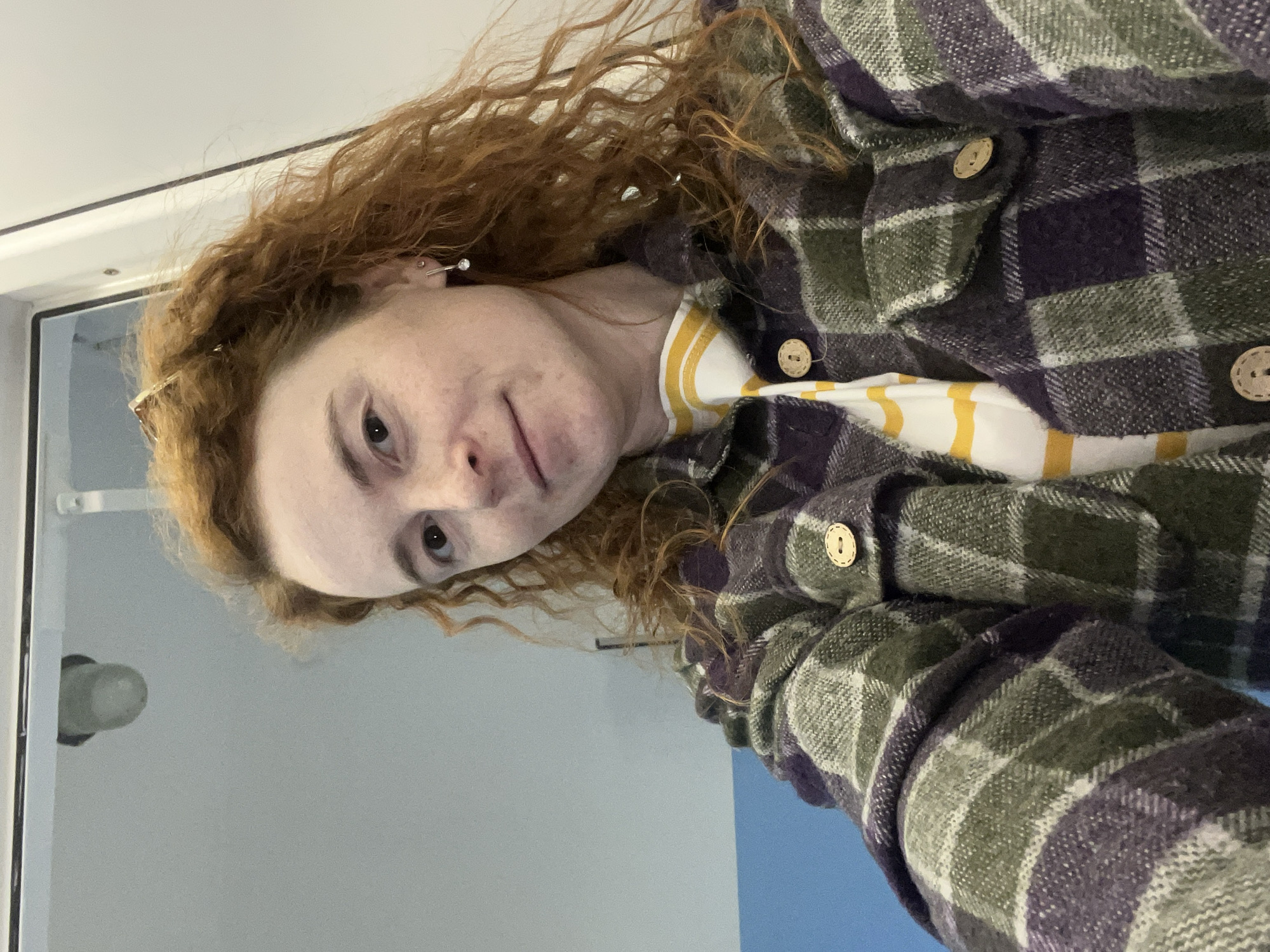 A woman with red curly hair, fair skin with freckles, and pierced ears is seen from a close, angled perspective. She is wearing a buttoned, plaid flannel shirt in shades of green and purple, with light brown buttons. A striped garment with yellow and white stripes is visible underneath the flannel. The background appears to be a room with a white wall near a doorway or opening. A light fixture is visible on the left. No information suggests a location in Chaikovsky, Russia.