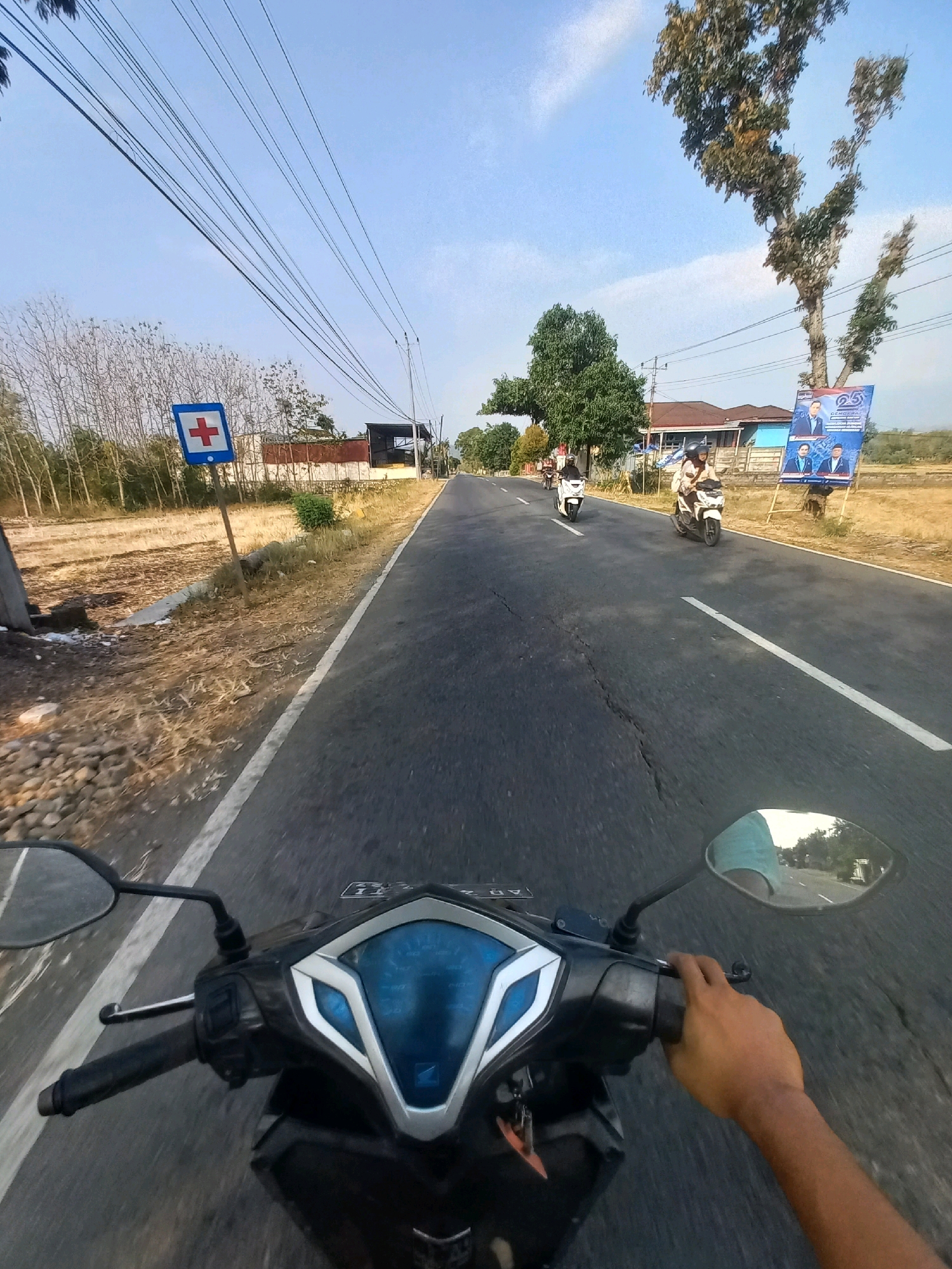 The image captures a first-person perspective of someone riding a motorcycle on a paved road. The road appears to be in a rural or semi-urban area, with dry, grassy fields and sparse trees lining one side. Overhead, a network of power lines stretches across the sky.
To the left of the road, a blue sign with a red cross is visible, indicating a medical facility or emergency service. Further down the road, there are a few buildings, including what looks like a residential structure with a reddish-brown roof and a larger building with a dark roof. On the right side of the road, a tall, leafy tree stands near a large, vertical banner featuring portraits of two men and text that appears to be political in nature.
Several other motorcycles are on the road ahead. Two motorcycles are closer to the foreground, with riders and passengers visible. One motorcycle, in the middle distance, has two people on it, and another motorcycle is further ahead with a single rider. The sky is clear and blue with a few wispy clouds. The overall impression is a snapshot of daily life on a road in a relatively undeveloped area, possibly in the city of Baturetno, Indonesia, given the context provided. The image shows a clear, sunny day.