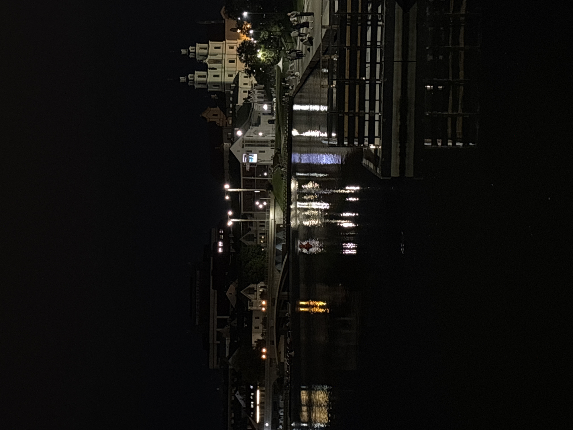 The image presents a nighttime panoramic view of the Svislach River in Minsk, Belarus, featuring illuminated architecture and reflections. The lower half of the frame is dominated by the dark water of the river, which exhibits prominent, elongated vertical reflections of artificial lights from the city.
On the right bank, the brightly lit **Holy Spirit Cathedral** stands out with its distinctive white Baroque facade and two prominent towers, each topped with a dark, pointed spire. Adjacent to the cathedral are other illuminated buildings, some displaying architectural detail. A paved promenade runs along this bank, where multiple indistinct groups of people are visible standing, walking, and interacting. Streetlights uniformly illuminate the promenade.
On the left bank, a multi-arched bridge with bright yellow/orange streetlights spans the river. Behind the bridge, a cluster of buildings with illuminated windows is visible, extending into the background. A larger, dark, multi-story building, possibly a modern complex, is silhouetted against the dark sky in the far background above the left bank.
On the river itself, near the bridge, several small boats or kayaks are moored. Further to the right on the water, a red-and-white striped cylindrical buoy is present. In the immediate lower right foreground, a dark wooden or metallic dock structure is partially visible, with its reflection clearly rendered in the water. The sky above is uniformly dark and devoid of visible stars.