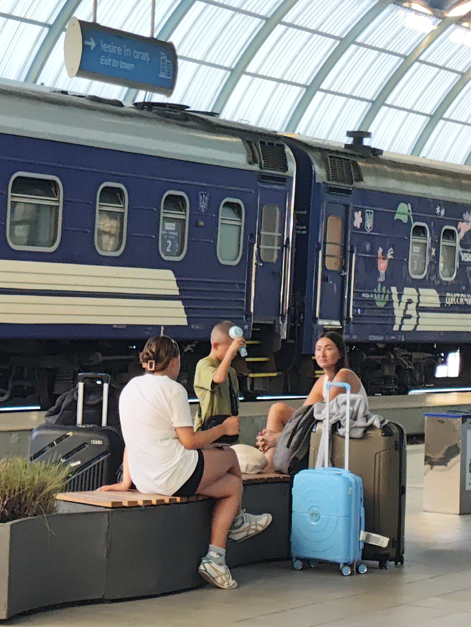 At a train station in Chișinău, Moldova, three individuals are seen waiting on a platform. A blue train with a gray stripe and white trim is parked at the platform. A blue sign with white text indicating "Exit to town" and Romanian text is hanging above the train. In the foreground, a woman with dark hair is seated on a bench next to a small blue suitcase and a larger brown suitcase. To her left, a child in a green shirt is standing and holding a small bottle. Further to the left, another person wearing a white shirt and black shorts is sitting on the bench with their back to the camera, their legs crossed. Luggage is visible near the bench, including a black rolling suitcase. The environment appears to be an indoor or covered train station with a high, arched ceiling.