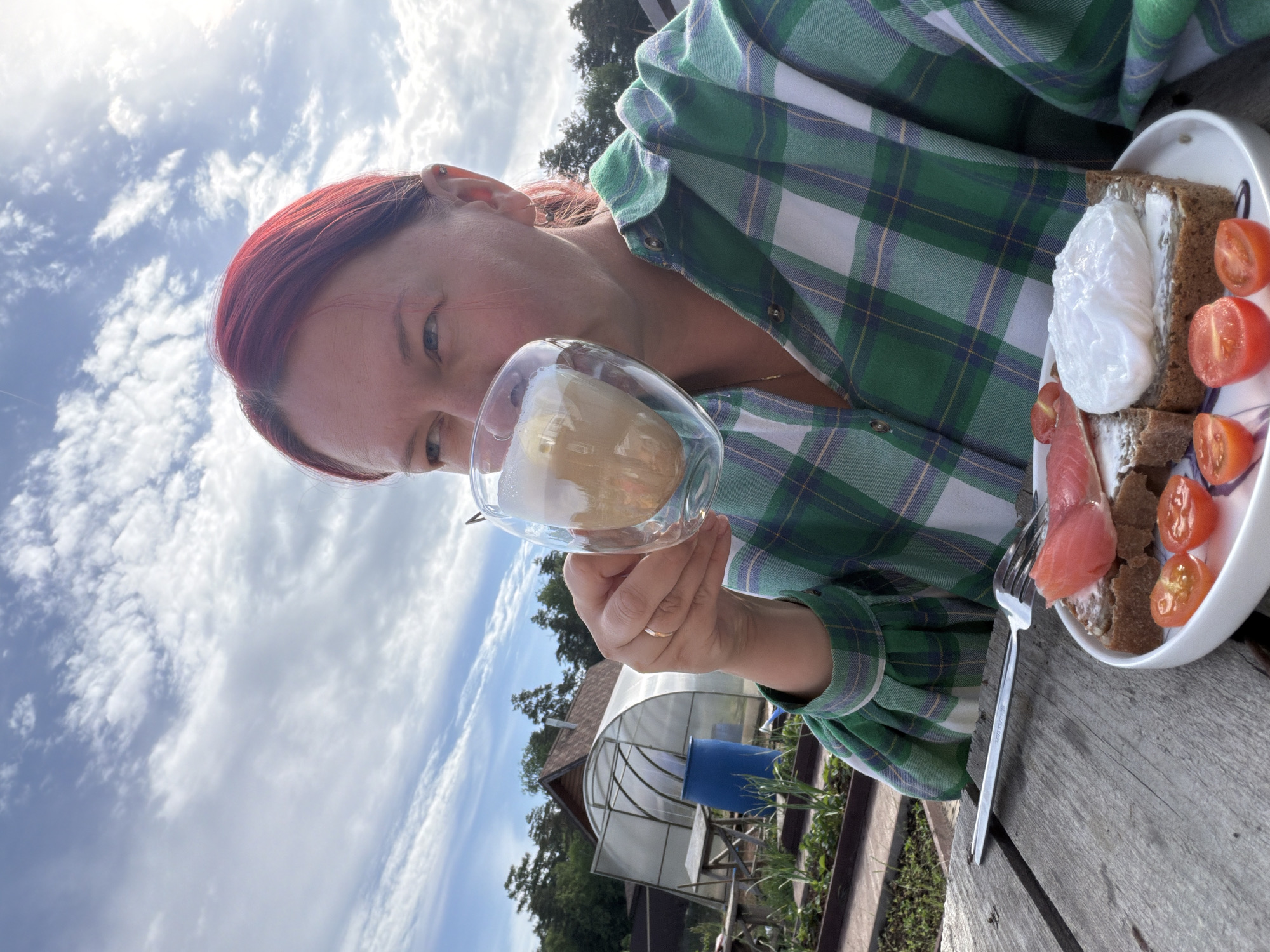 A person with red hair is holding a glass of liquid up to their mouth, presumably to drink. They are wearing a green and white plaid shirt. To their right is a plate with a slice of bread topped with a poached egg, smoked salmon, and sliced cherry tomatoes. There is a fork on the wooden surface next to the plate. In the background, the sky is visible with scattered clouds. A greenhouse and a small wooden structure are also visible in the background. Based on geolocation metadata, the photo was taken in Chaikovsky, Russia.