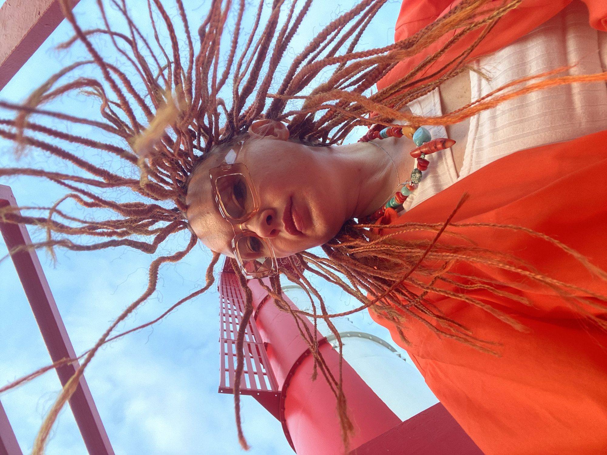 This media file is a close-up, low-angle, upward-facing shot of a person with dreadlocks. The subject is wearing large, square, tinted glasses and a colorful beaded necklace. They are also wearing an orange garment, possibly a dress or shawl, and a white textured top underneath. The shot captures the person from the chest up, with their head tilted back and their dreadlocks spread out against a bright blue sky with some clouds.
In the background, there is a red structure that appears to be part of a slide or a similar amusement park feature, suggesting a location like an amusement park or a water park. The lighting suggests it is daytime, likely sunny given the brightness of the sky. The perspective makes the person and the red structure appear very large.
The overall impression is one of a playful and perhaps adventurous moment, with the subject looking up towards the sky. The vibrant colors of the clothing and necklace contrast with the blue sky and red structure. No visible text is present in the image. The context provided indicates the location is Sochi, Russia, which further supports the idea of an outdoor leisure or entertainment setting.
