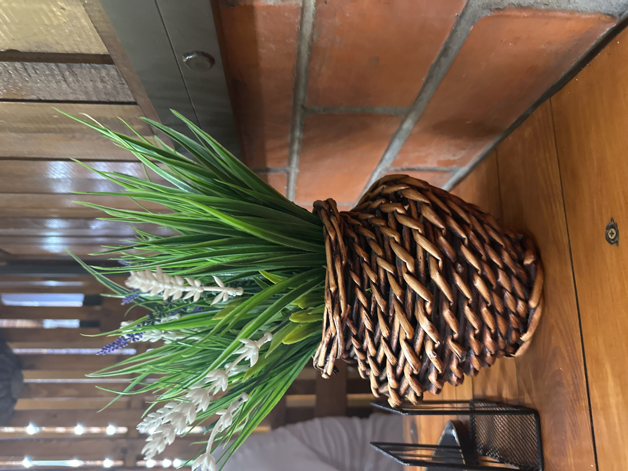 This is a close-up, medium shot of a woven basket holding faux greenery and flowers, positioned on a wooden surface. The basket is a rich brown, intricately woven with a tapered shape that widens towards the top. Inside, several stalks of bright green grass-like foliage are prominent, interspersed with white, feather-like blooms and some small clusters of purple flowers.
To the left of the basket, a black metal desk organizer is visible. The background consists of a brick wall with orange-toned bricks and grey mortar, and further back, a slatted wooden structure. A dark metallic band runs horizontally above the brick wall. The lighting suggests an indoor environment, possibly a cafe or restaurant, given the rustic materials and table setting. No people are clearly visible, though a blurred figure in a white shirt is faintly discernible in the far left background. The image appears to be taken at eye level. No specific location in Bol'shoi Kichmai, Russia is identifiable from the visual information presented.