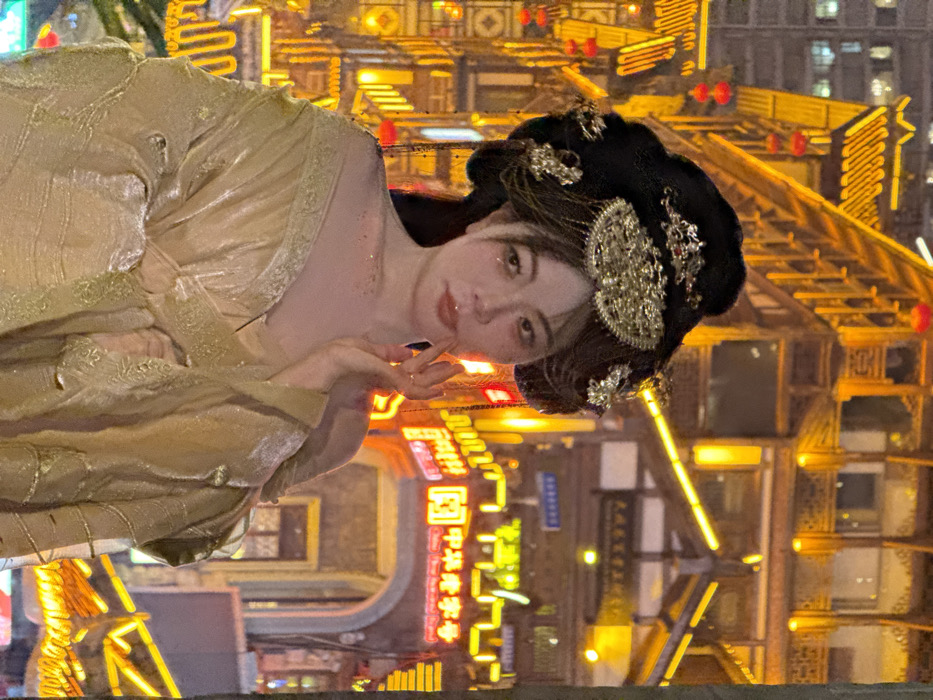 The image captures a female individual, likely a young adult, in a posed upright position against an illuminated traditional Chinese architectural backdrop at night. The location is identified as Hongyadong Commercial Street in Yuzhong District, Chongqing, China.
The subject wears an elaborate black updo hairstyle adorned with multiple gold-colored, intricate metal hairpins and a prominent fan-shaped headpiece. Light-colored strands frame her face. Her makeup includes visible reddish blush, eye makeup, and glossy lips. Subtle glitter particles are present on her left shoulder and upper chest. She is dressed in a flowing, layered garment of a shimmering champagne or golden color, consistent with traditional Chinese Hanfu, featuring textured or brocaded patterns. Her right hand is raised, with fingers delicately touching her chin or lower lip. She gazes directly forward.
The background consists of multi-story buildings exhibiting traditional Chinese architectural elements, including curved roofs, wooden balconies, and intricate eaves. These structures are extensively outlined and lit with warm, golden artificial lights, creating a vibrant nocturnal scene. Red lanterns hang from several buildings. In the mid-right, a dark archway is visible, above which a prominent red neon sign with white Chinese characters reads "中华美食号" (Chinese Delicacy Number). Additional illuminated signs with Chinese characters are present on adjacent buildings.