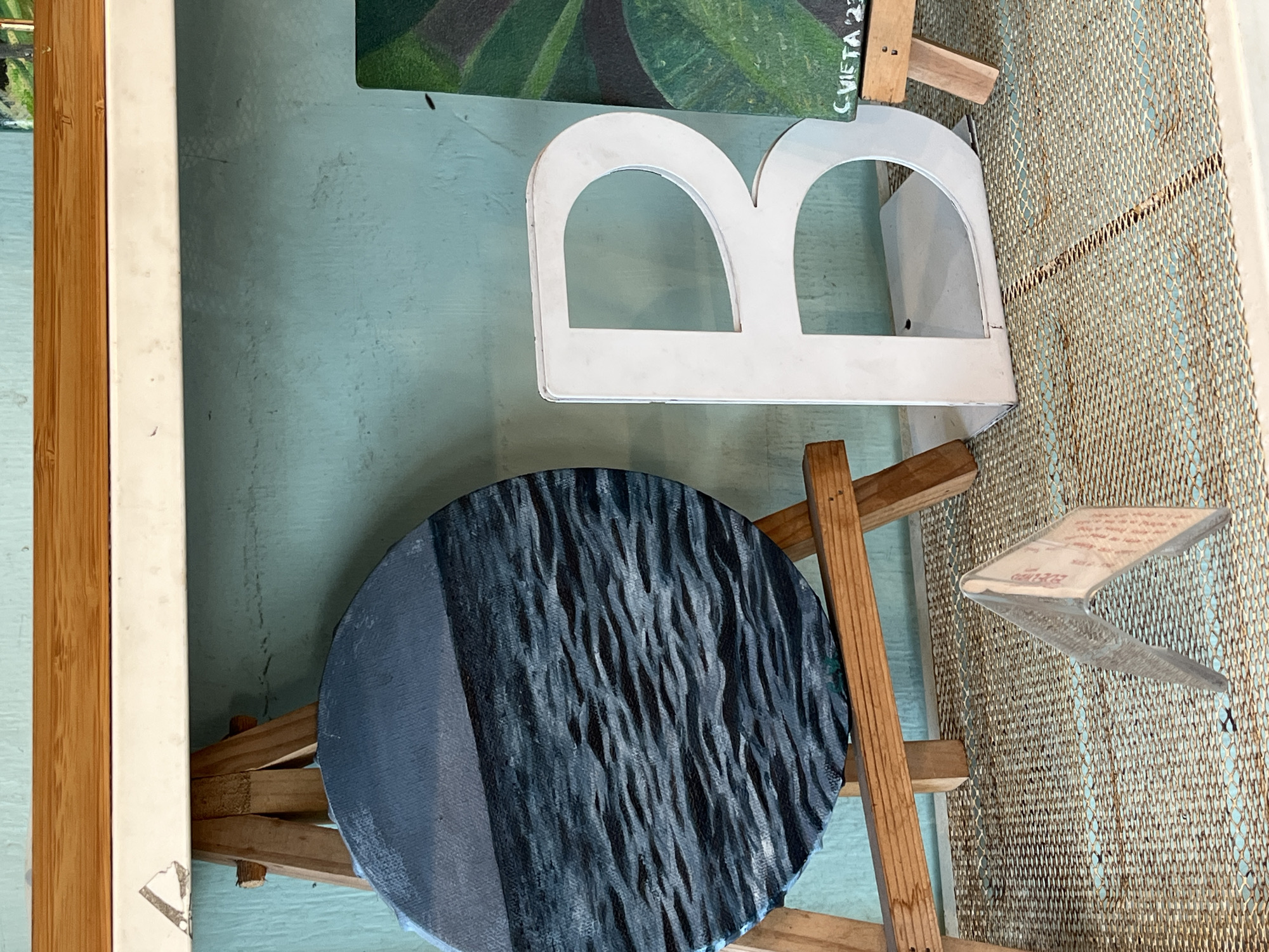 The image shows an interior space with a light blue wall. Several objects are present, including a framed picture with a wooden and white frame, a green painting with illegible writing, a white letter "B", a round painting with a textured black and gray surface, a wooden easel, and a piece of paper affixed to a beige woven surface. No people are visible. The location is listed as Unknown, Indonesia, but there's no additional information to confirm this.
