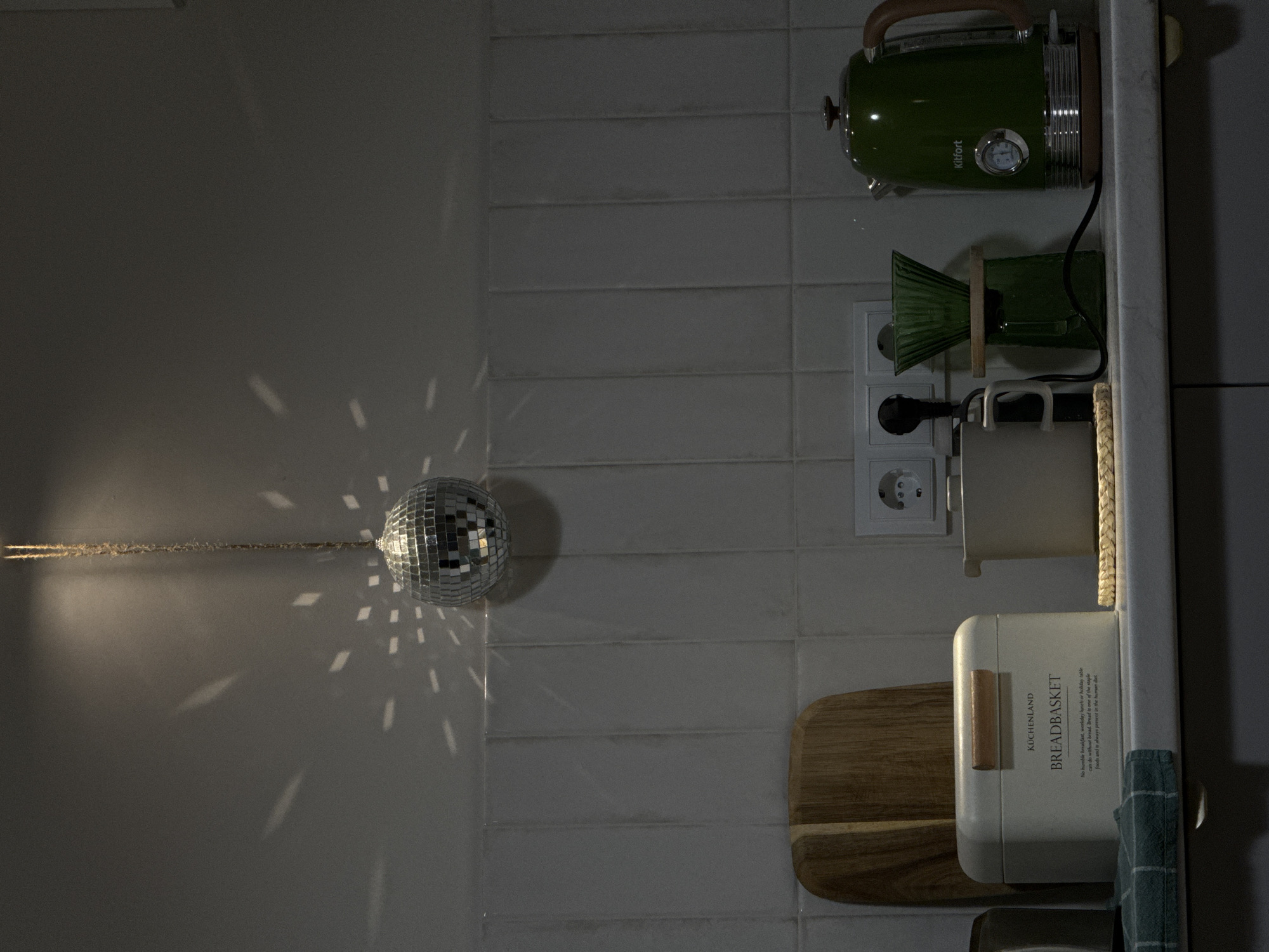 The image displays a dimly lit indoor scene, likely a kitchen, featuring a combination of functional appliances and decorative elements. The photograph is oriented vertically, showcasing a tiled wall on the left and a kitchen counter on the right.
A small, reflective disco ball is suspended on the light-colored wall, projecting numerous rectangular patterns of light onto the surrounding surface, suggesting an external light source is illuminating it. The wall behind the disco ball and along the counter is covered with light-grey or white horizontal subway tiles.
On the kitchen counter, various items are arranged. At the top of the frame, a distinct green electric kettle, adorned with wooden accents and a visible thermometer display, proudly bears the "Kitfort" brand. Below the kettle, a green, ridged glass item, possibly a pour-over coffee carafe or a decorative vase, is perched on a small wooden stand. An electrical outlet with a plugged-in cord is visible on the tiled wall directly behind these items. Further down the counter, a light-colored container with a handle sits beside a white bread box. The bread box is branded "KÜCHENLAND" and labeled "BREADBASKET," with additional text stating, "No humble breakfast, weekday lunch or holiday table can do without bread. Bread is one of the staple foods and is always present in the human diet." A wooden cutting board leans against the tiled wall behind the bread box, and a green checkered towel hangs from the counter's edge.
The scene appears to be set indoors, likely during the evening or night, given the low ambient light and the prominent light reflections from the disco ball. No individuals are present, and no particular activity is occurring, presenting a static view of a well-equipped kitchen space.