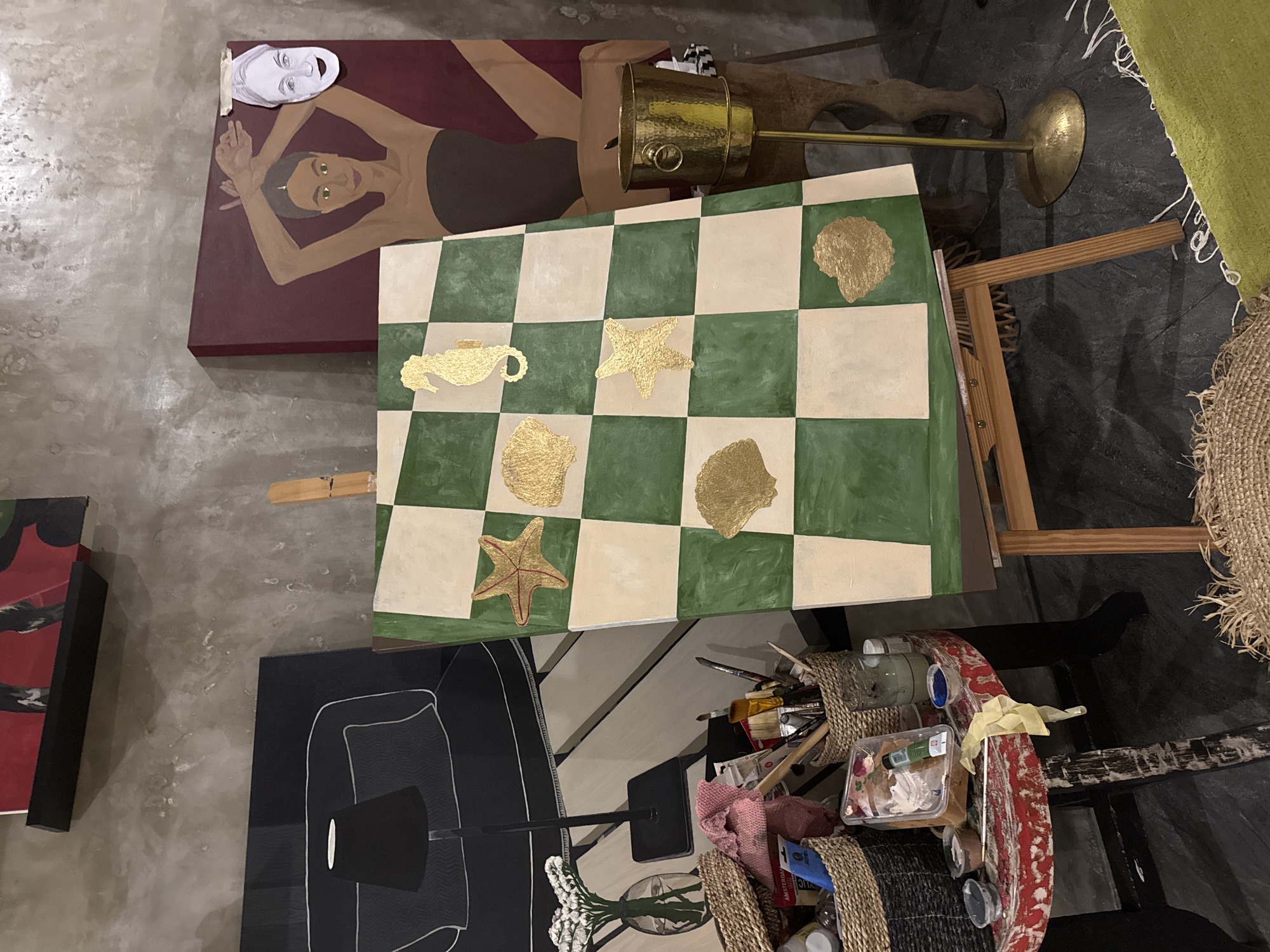 This indoor scene appears to be a workspace in North Kuta, Indonesia, filled with art and artistic materials. Several paintings are visible, including one depicting a reclining woman with a mask lying near her head, another of a black and white design, and a chessboard-patterned piece adorned with golden figures of sea creatures. An adjustable brass floor lamp stands near the paintings. The area also contains a cart holding paintbrushes, containers, and other art supplies, along with baskets holding various items. An easel is present supporting one of the paintings. The setting suggests an artist's studio or a creative space.