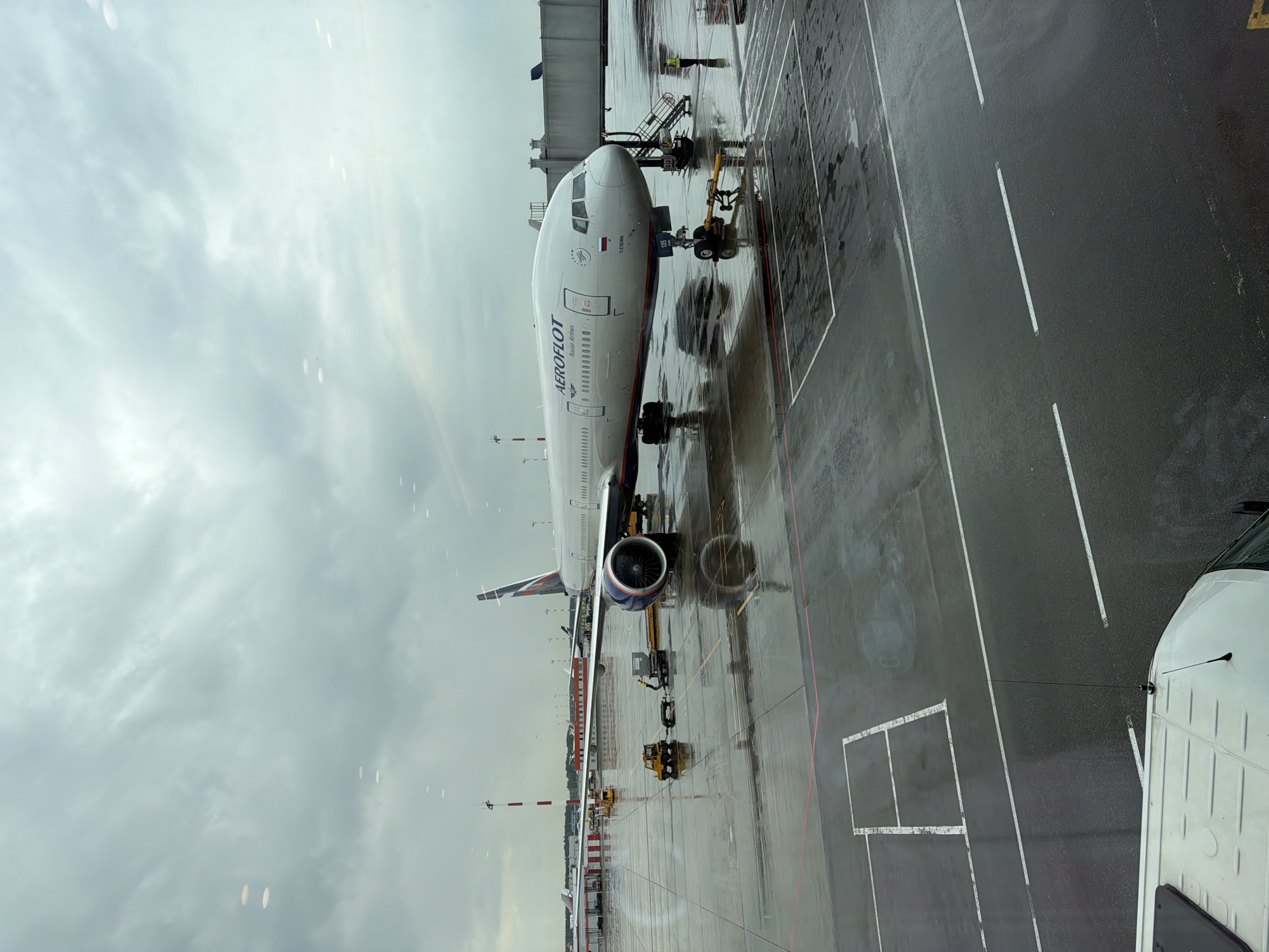 An Aeroflot airplane, identified by the "AEROFLOT Russian Airlines" livery and the Aeroflot logo on its tail, is parked on a wet tarmac at an airport. The plane is a large passenger jet with visible engines and landing gear. Several ground vehicles are present near the aircraft, including a yellow tow tractor with a cargo cart and a white van in the foreground. A jet bridge is connected to the aircraft. A figure in a yellow safety vest is visible on the tarmac near the jet bridge. The airport tarmac is marked with painted lines and appears wet, suggesting recent rain. In the background, other aircraft and airport infrastructure, including a control tower with red and white markings, are visible. The sky is overcast and cloudy. The scene is likely Sheremetyevo International Airport (SVO) in Khimki, Russia, given Aeroflot's status as the flag carrier of Russia and its primary hub at Sheremetyevo.