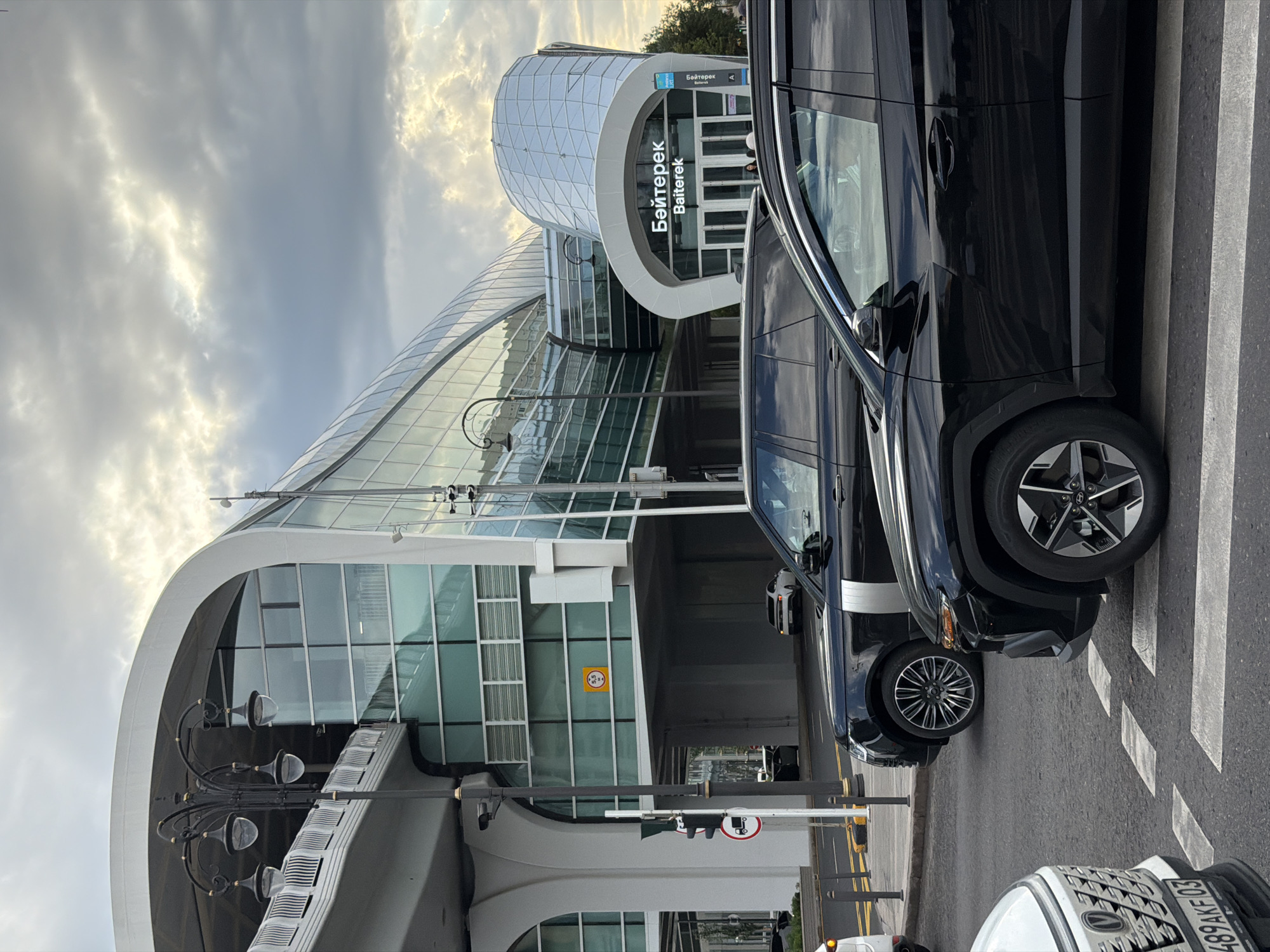 The image depicts the Bayterek tower in Astana, Kazakhstan, under a cloudy sky. In the foreground, several black SUVs are parked on a street, with a white vehicle partially visible to the left. The Bayterek tower is a prominent landmark, featuring a modern architectural design with glass and white curved structures. A sign above the entrance reads "Байтерек Baiterek". Traffic lights and street lamps are also visible. The scene appears to be an urban environment with a mix of modern architecture and vehicular traffic.