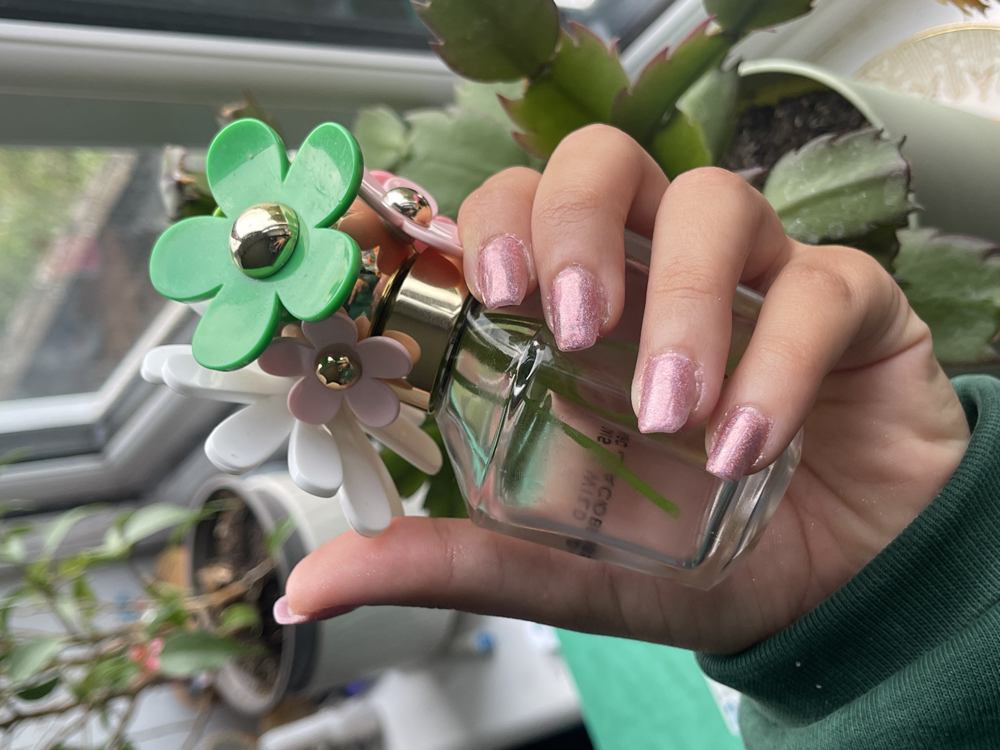 A person holds a bottle of Marc Jacobs Daisy Wild Eau de Parfum. The bottle's cap is decorated with a green daisy. The person has pink glitter nail polish. The background includes a window, potted plants, and a green sweater. The location is not discernible from the image.