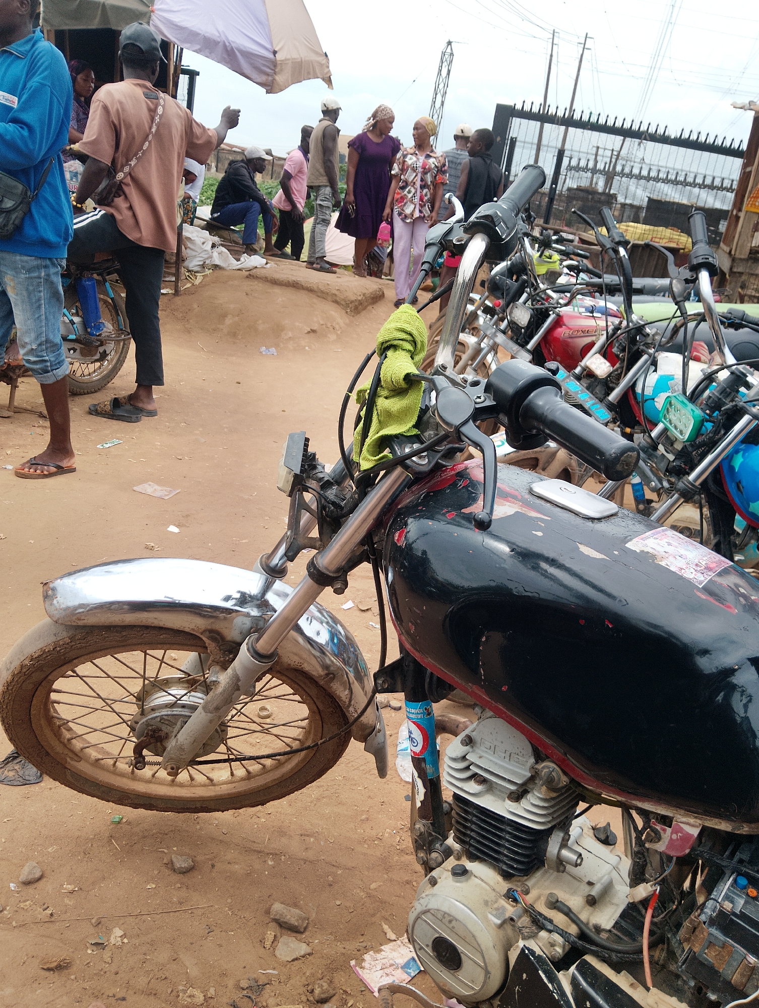 In the city of Ijoko, Nigeria, a scene unfolds with several motorcycles parked and people standing around. A man in a blue jacket and jeans stands to the left, his attention focused elsewhere. Next to him, a person wearing a tan shirt and black pants is seated on a motorcycle, with a patterned strap across their chest.
Behind them, a group of people are visible, some interacting while others stand by. A woman in a purple dress and a woman in a patterned shirt and pink pants are seen further back. Several other individuals are scattered throughout the background.
The foreground is dominated by a black motorcycle, its chrome fender and wheel prominent. A lime green cloth is draped over the handlebars. The motorcycle appears weathered, with some scratches and stickers on its fuel tank. Other motorcycles are parked closely together behind it, their handlebars and various parts creating a cluttered yet organized arrangement. The ground is made of dry, dusty earth with some scattered debris. In the background, a metal fence and some electrical poles are visible, suggesting an urban or semi-urban setting. The sky is overcast, indicating a cloudy day.