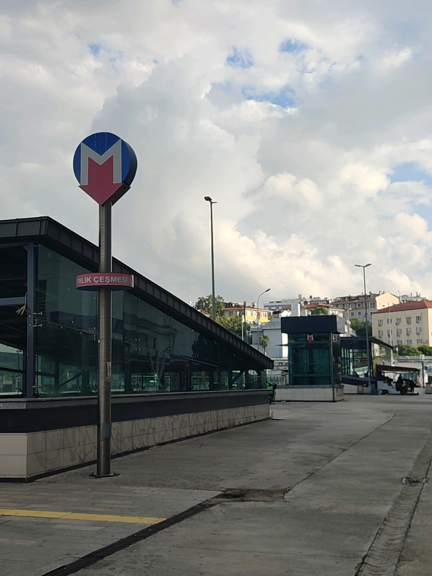 The image shows a street-level view of a metro station entrance in Kadıköy, Turkey. A prominent blue and red metro sign with a white "M" is visible on a pole to the left. Below the "M" sign, a circular red sign reads "Ri̇lik Çeşmesi" in white lettering, which appears to be a station name. The entrance itself is a modern structure with a dark-colored roof and glass walls, reflecting the cloudy sky. The ground in front of the station is paved with concrete, and a yellow tactile paving strip is present on the left side of the walkway. In the background, several buildings with various architectural styles and colors are visible, characteristic of an urban environment. A street light stands to the right of the metro entrance, and further back, another metro station entrance or building with a similar "M" logo can be seen. There are a few people and vehicles in the distance near the right side of the frame, suggesting ongoing activity. The overall scene depicts a public transportation hub within a city.