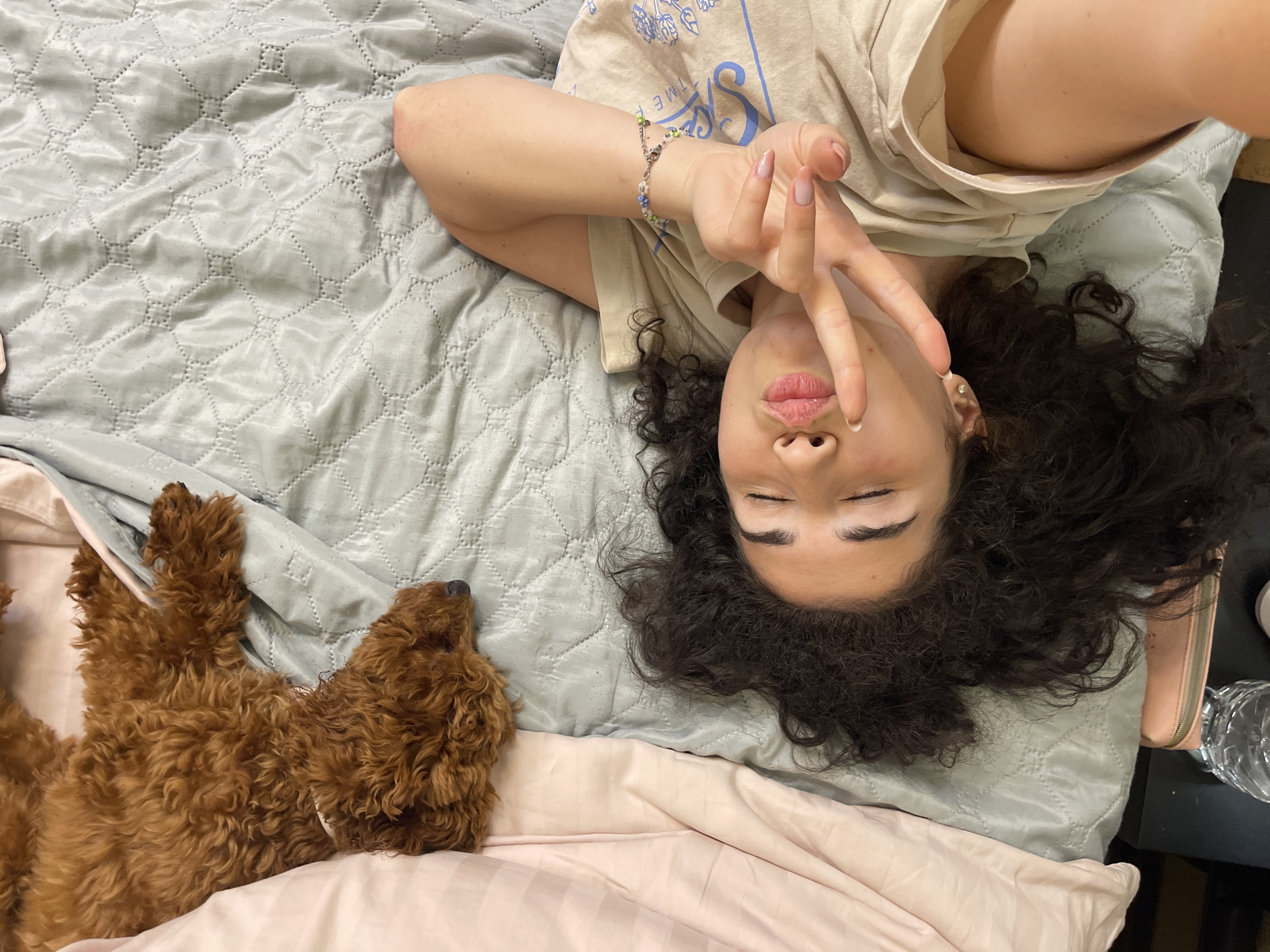 A medium-shot, high-angle, top-down selfie shows a young woman with dark, curly hair lying on a bed next to a golden doodle dog. The woman is wearing a tan t-shirt and has her eyes closed with her lips pursed. She is making a peace sign with her right hand, which is positioned near her face. Her left arm is resting behind her head. A beaded bracelet with green and blue beads is visible on her left wrist. The bed is covered with a light blue quilted comforter and a pink duvet is to the left of the frame. A small pink wallet and a partially filled water bottle are visible on a dark surface to the bottom right of the image. The scene appears to be an indoor setting, likely a bedroom. The image does not provide any information that would allow for the localization of the scene in Sofia, Bulgaria.