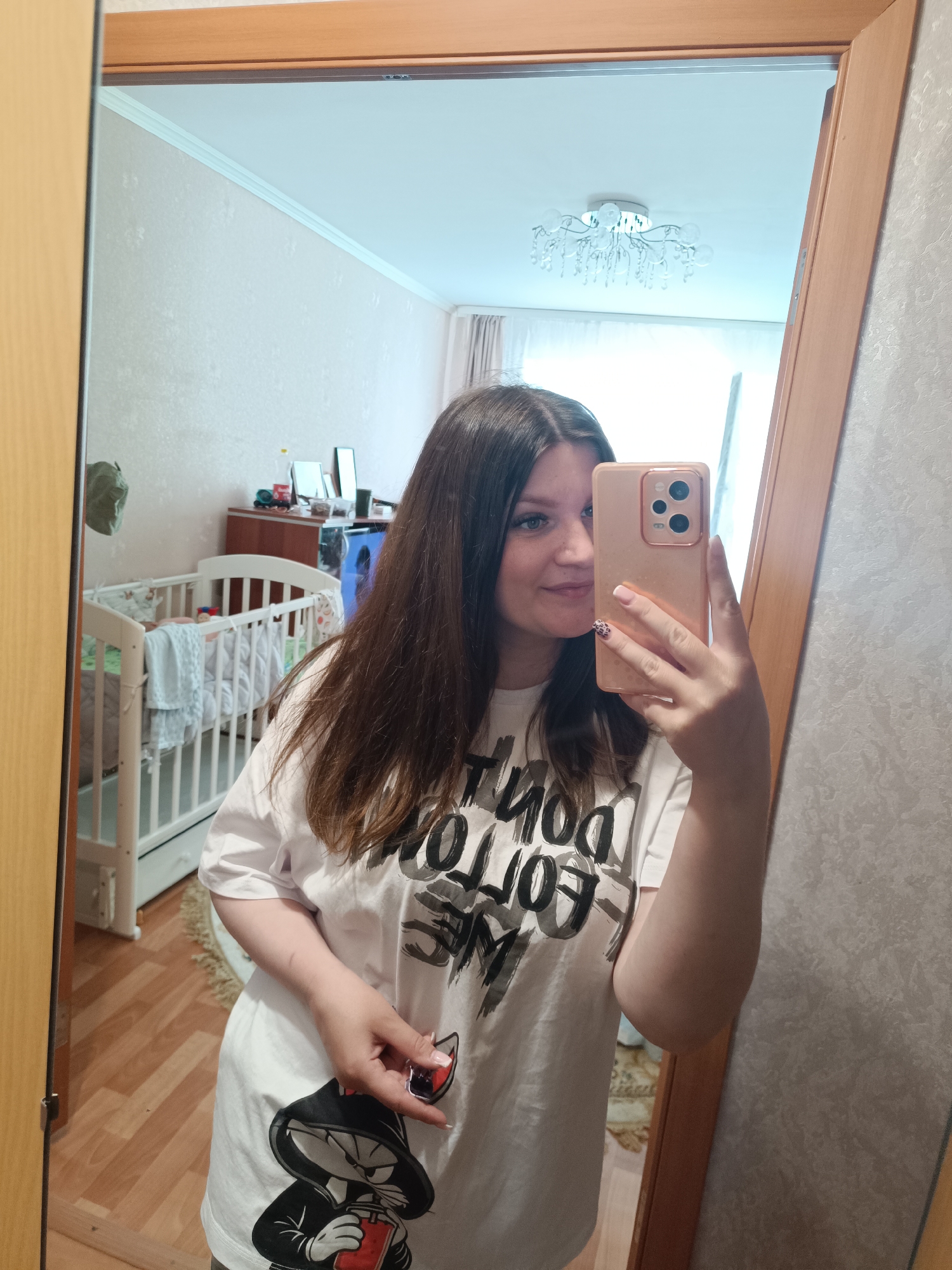 A young woman is taking a selfie in a mirror. She is wearing a white t-shirt with black graphics. She is holding a pink phone. Behind her in the mirror is a room with a crib, a dresser, and a chandelier. The reflection in the mirror shows the inside of an apartment in Tobolsk, Russia.