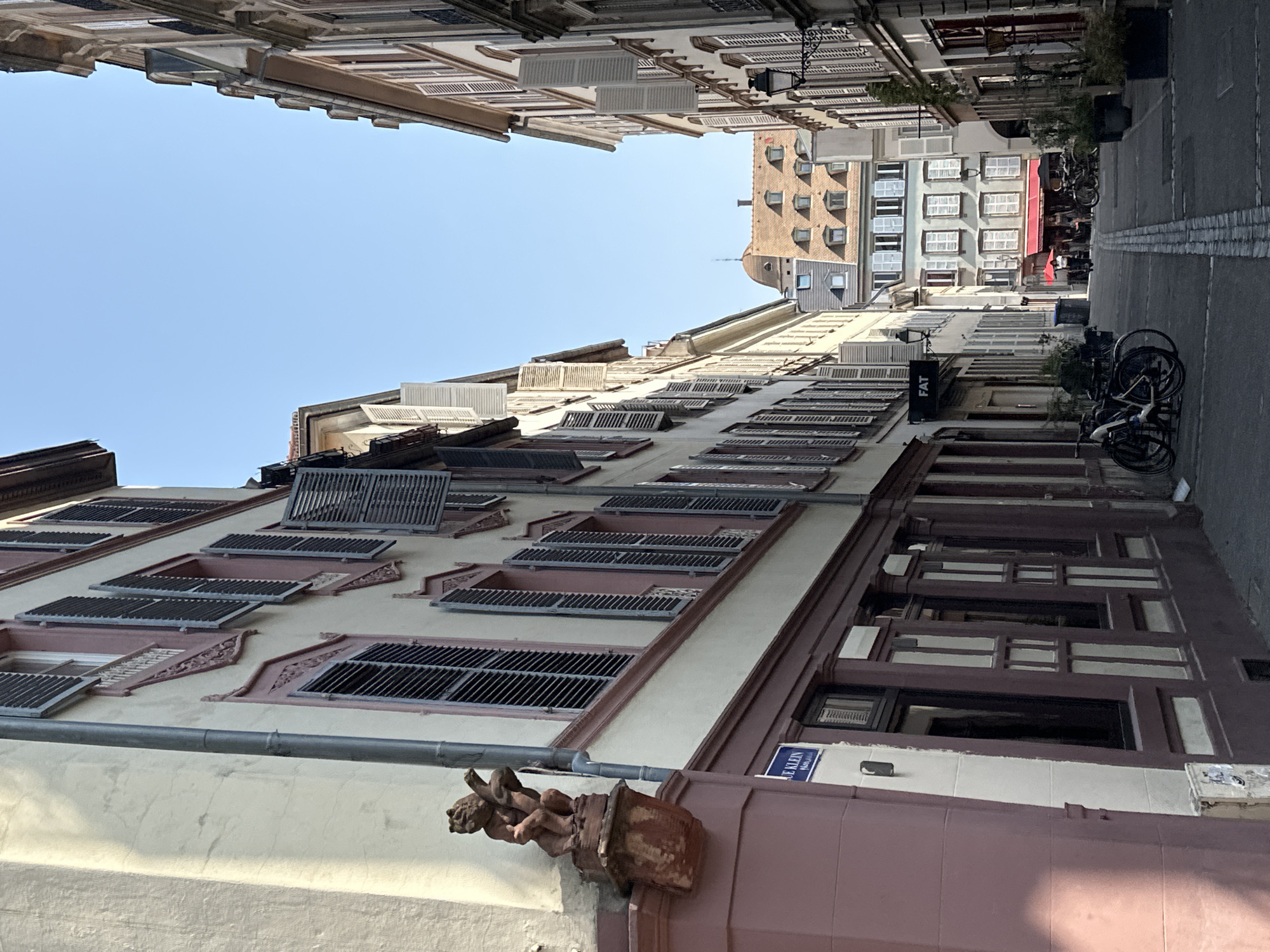 The image depicts a narrow, cobblestone street in Strasbourg, France, likely in the Petite France district, lined with historic, narrow buildings. The facades of the buildings are painted in pale yellow and rose colors, with dark wooden beams and shutters on the windows. A small, terracotta statue of a cherubic figure is visible on the corner of one building. The sky is clear and blue. In the background, several people are seen walking and standing outside a cafe with a red awning. Bicycles are parked along the side of the street. A street lamp is visible on the right side of the image. The overall atmosphere is tranquil and picturesque, characteristic of a European old town.