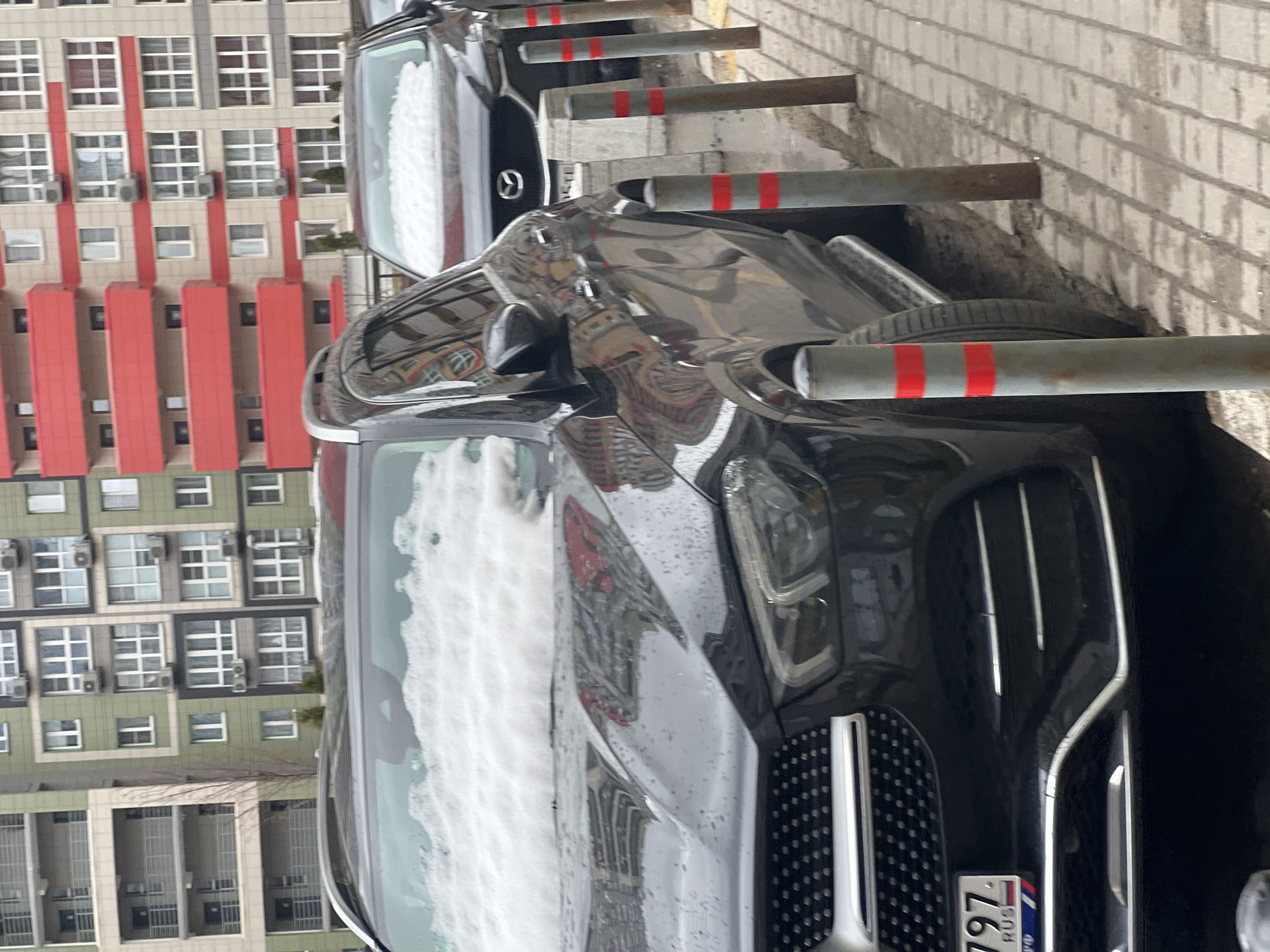 The image captures an urban street or parking area in Khimki, Russia, featuring two parked cars, residential buildings, and a row of bollards.
In the foreground, a dark-colored SUV, likely a Mercedes-Benz, is prominently displayed. Its large front grille with vertical chrome slats and intricate headlights are clearly visible. The hood and windshield of the SUV are partially covered with a layer of snow or frost, suggesting cold weather. The glossy surface of the car reflects parts of the surrounding buildings and poles. Behind this SUV, a dark sedan is parked, with a Mazda logo visible on its front fender.
A series of robust gray metal bollards, each marked with two distinct red horizontal stripes, line the edge of the parking area, separating it from what appears to be a sidewalk or another section of the street. These bollards show signs of wear and minor rust.
In the background, multi-story residential buildings form the backdrop. One building has a light beige or tan facade with striking vertical red panels, numerous windows, and multiple external air conditioning units. Another building features a greenish-grey facade with different window configurations and more air conditioning units, some sections appearing to have balconies or recessed areas.
The scene takes place on an overcast or cloudy day, given the diffused light and lack of strong shadows. The presence of snow/frost on the SUV and the wet appearance of the car bodies and ground indicate cold and damp conditions.
On the front license plate of the SUV, the partial text "797 RUS" is visible, confirming its Russian registration. The overall impression is a typical urban parking scene during a chilly, possibly post-snowfall, day.