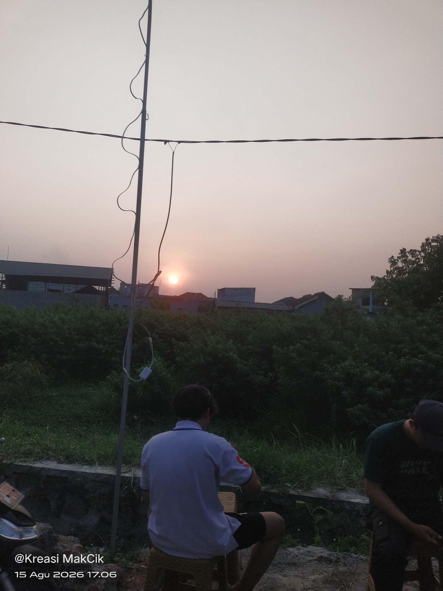 The image depicts an outdoor scene at dusk, possibly in Gilang, Indonesia. The sun is setting, casting a soft glow on the sky. In the foreground, two individuals are visible. One person, wearing a light blue polo shirt and dark shorts, is seated on a stool facing away from the camera, towards the sunset. The other person, wearing a dark t-shirt and a cap, is partially visible on the right, also facing away. A motorcycle is parked on the left side of the frame. Behind the people, a dense green bush line forms a backdrop, with a stone wall running horizontally. In the mid-ground, various buildings and structures are silhouetted against the sky. A tall pole with wires extends vertically through the center of the image. The overall atmosphere is calm and serene, with the natural light of the sunset dominating the scene.