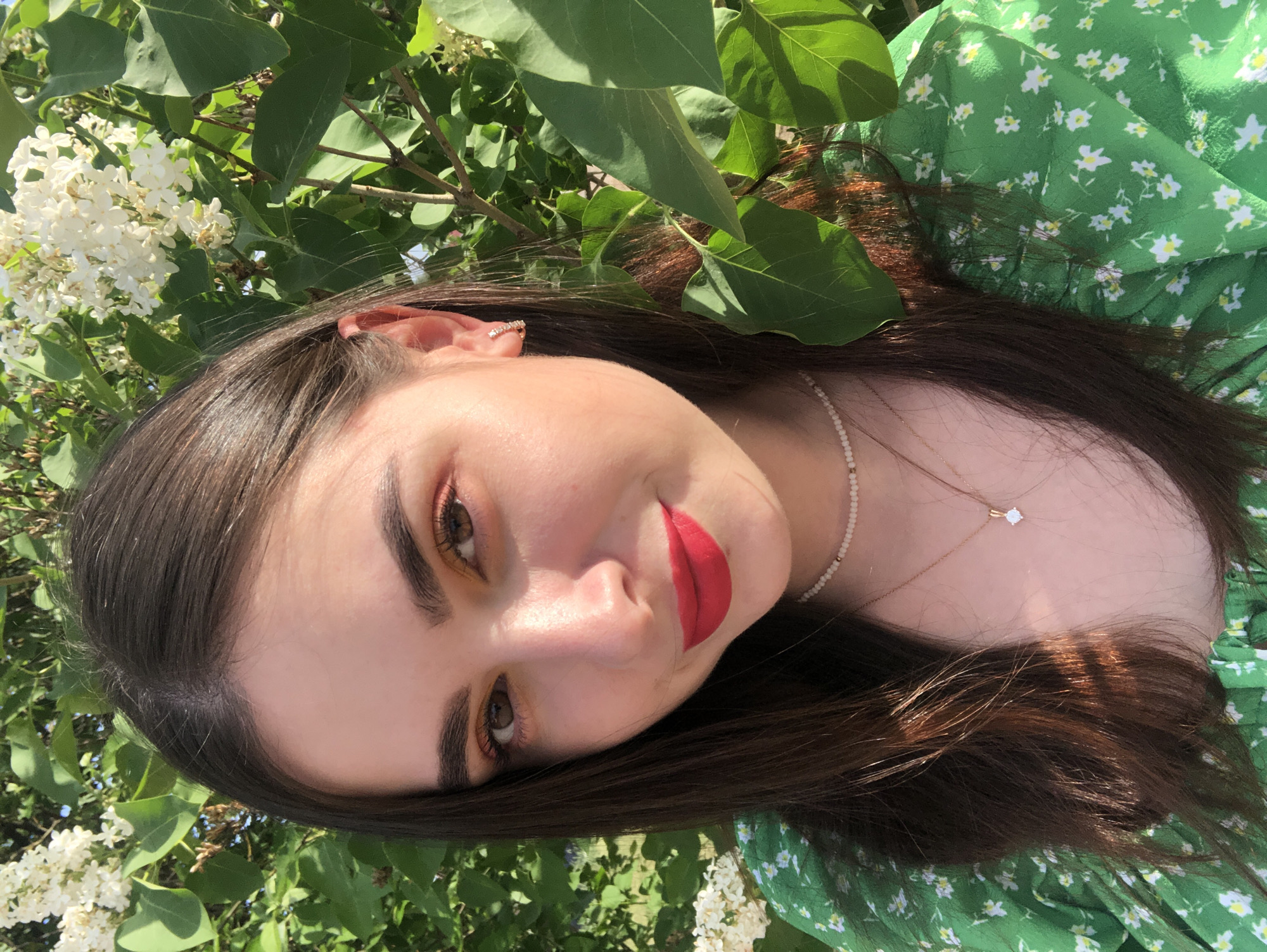 A young woman is facing the camera, surrounded by leafy green foliage and white flowers, likely lilac. She has long brown hair, fair skin, and is wearing red lipstick. She is wearing a green floral dress and a necklace. The photo appears to be taken outdoors in natural light. It is impossible to determine if the location is Noviy, Russia.