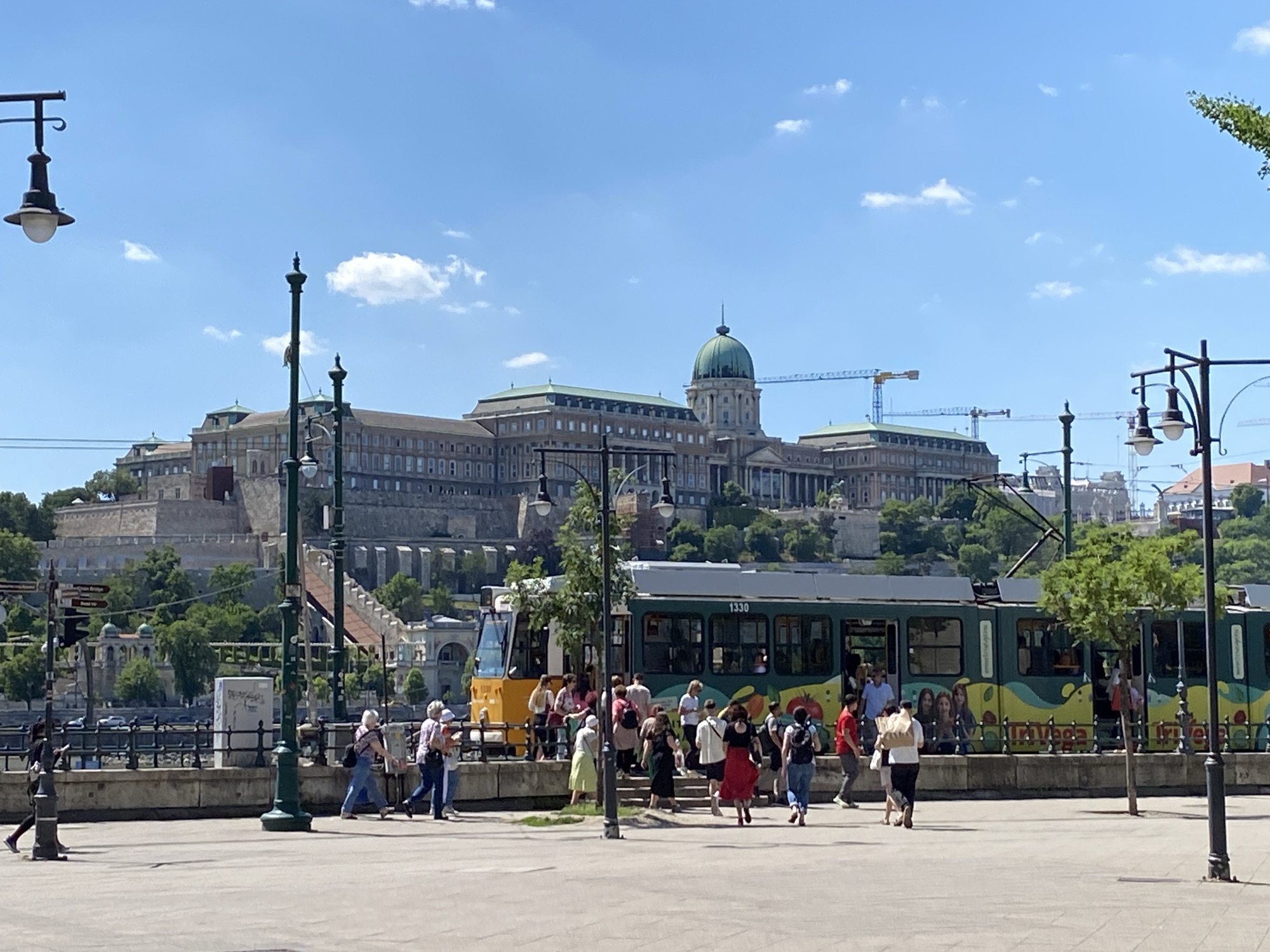 The image depicts a sunny scene in Budapest, Hungary, showing the Buda Castle in the background. In the foreground, a crowd of people are boarding or disembarking a yellow and green tram, numbered "1330" and decorated with colorful fruit and vegetable images. The tram runs along the riverfront. Several lamp posts are visible along the riverbank. The Buda Castle is a large, imposing building with a central dome. There are construction cranes present behind the castle. The sky is blue with scattered white clouds.