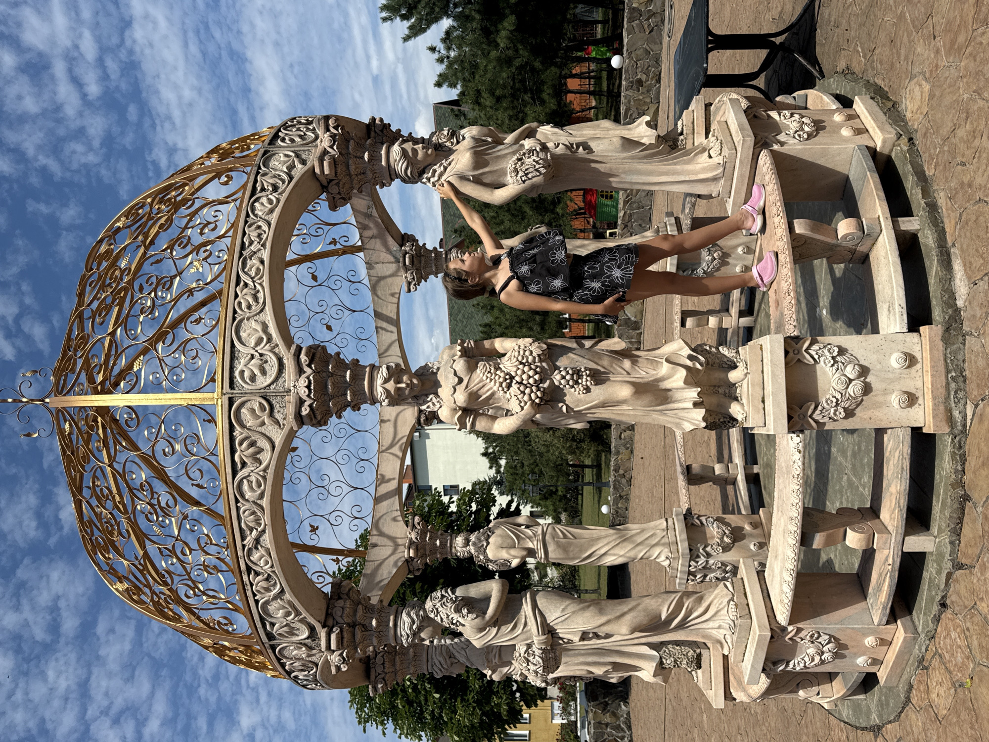 A young woman with dark hair, wearing a black and white floral dress and pink sandals, poses playfully in front of an ornate, classical-style fountain. The fountain is made of carved stone, featuring multiple statues of women holding grapes and bouquets, and is adorned with decorative floral motifs. A golden, wrought-iron canopy arches over the fountain, decorated with intricate scrollwork. The woman is standing on the edge of the fountain, reaching out to touch one of the statues.
The scene appears to be outdoors, bathed in the warm light of a sunny day. The sky above is a vibrant blue, with scattered white clouds. In the background, there are lush green trees, stone walls, and buildings, suggesting a park or garden setting. The overall mood is lighthearted and joyful, with the woman's posture and expression conveying a sense of fun. The time of day appears to be midday or early afternoon, given the bright sunlight and shadows. There is no visible text in the image.