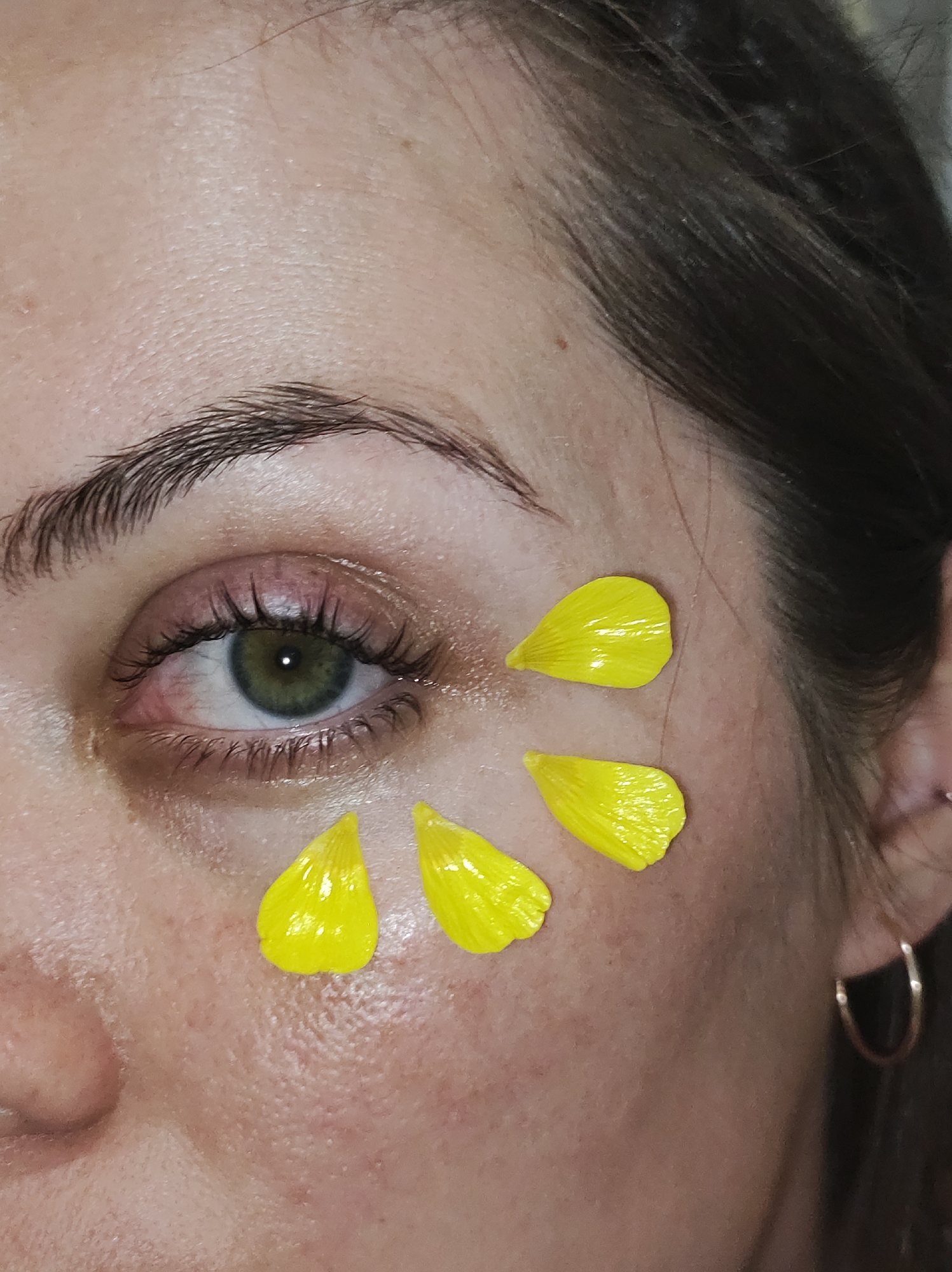 Close-up portrait of an individual's face, primarily focusing on the left eye and surrounding facial features. The subject exhibits a fair-to-medium skin tone with visible pores and minor epidermal irregularities. The left eye has a green iris and a central pupil; the sclera presents with mild erythema, particularly in the medial canthus region. Eyelashes are dark and appear enhanced. The eyebrow is well-defined and dark brown. Five bright yellow, glossy flower petals are adhered to the skin of the left infraorbital area and upper cheekbone. A simple, gold-toned hoop earring is visible in the subject's right earlobe, partially obscured by dark brown hair that frames the right side of the face. The scene is located in Unknown city, Unknown.