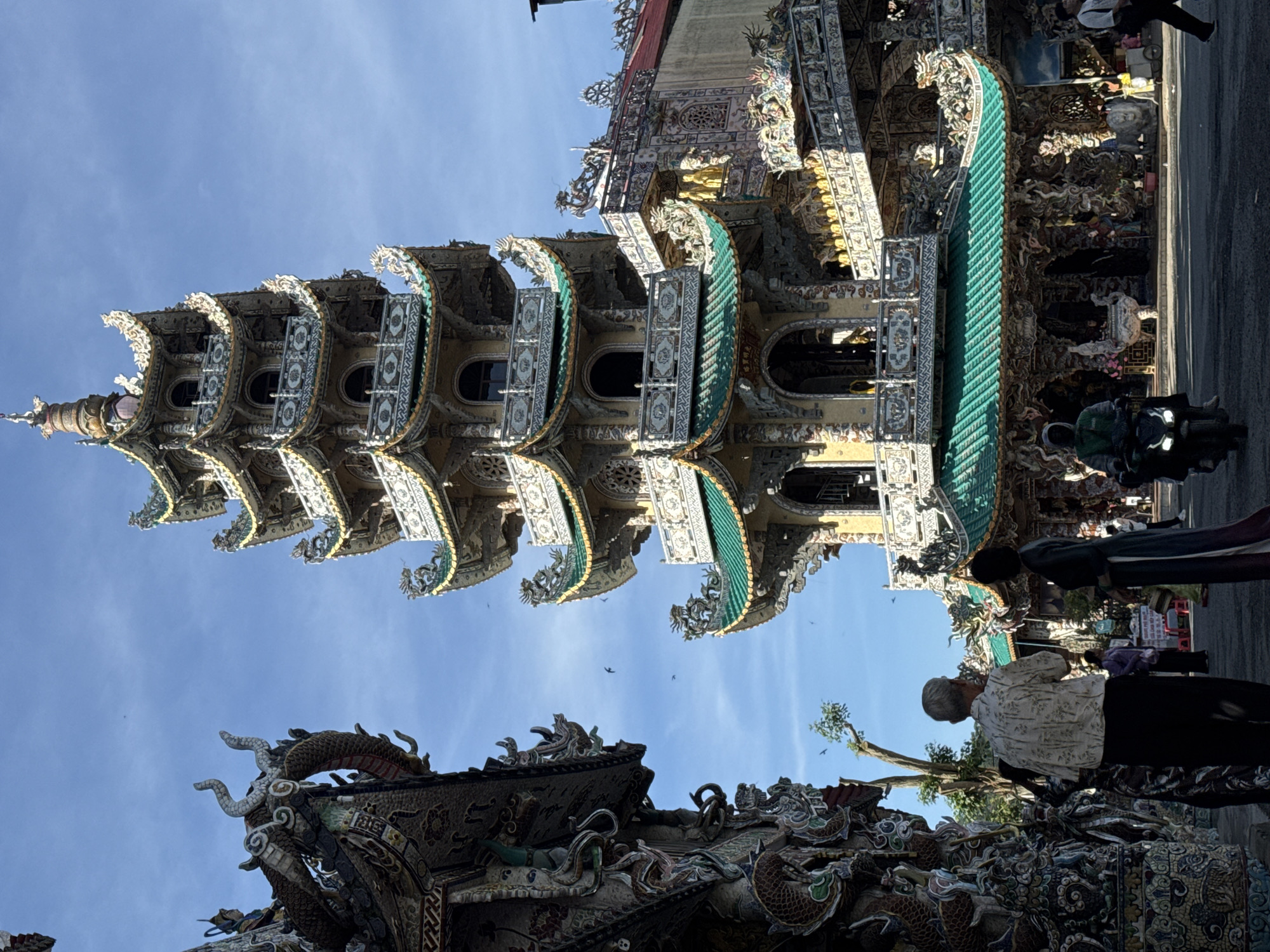 In the city of Xuân Thành, Vietnam, under a clear blue sky with scattered birds, a prominent, multi-tiered pagoda with green tiled roofs and intricate dragon and floral motifs dominates the scene. The pagoda's ornate facade features multiple levels with balconies and arched windows, decorated with colorful ceramic tiles and statues. To the left, another elaborately decorated structure, also adorned with dragon sculptures, partially obstructs the view of the sky. In the foreground, a man with gray hair and a patterned shirt stands facing away from the camera, next to a woman in a dark dress. Further into the scene, a person rides a scooter and another individual walks in the background near the pagoda's entrance. The ground is paved, and there are indications of other buildings and decorative elements associated with the temple complex.