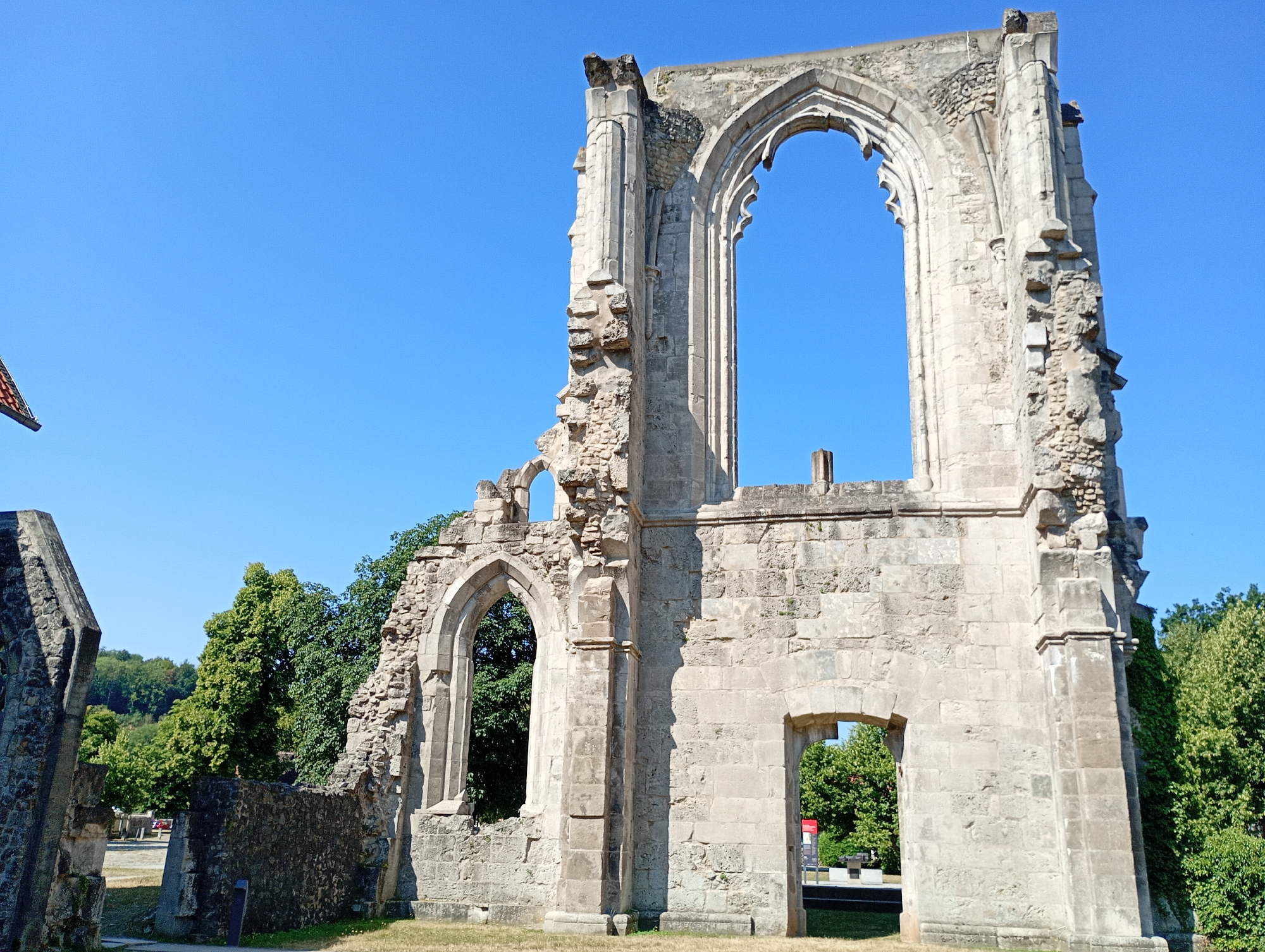 The image shows the ruins of the former Walkenried Abbey in Walkenried, Germany. The ruins are made of stone and have a Gothic architectural style, with large arched windows and weathered facades. The sky is clear and blue, and there are trees and greenery in the background, suggesting it is a sunny day. The ruins appear to be well-preserved but are clearly old and partially collapsed, indicative of their historical significance. There are no people visible in the image. The scene is one of historical preservation and natural surroundings.