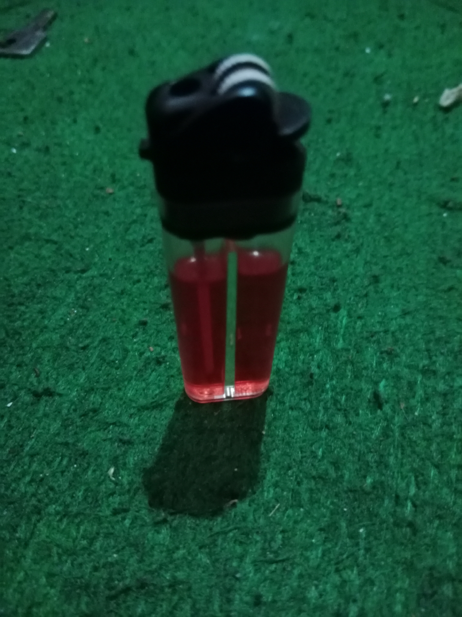 A transparent lighter with red fluid is lying on a green, carpet-like surface. The lighter has a black top with some silver accents. The background is a blurred green surface with some lighter specks. There are no people or animals visible in the image, nor are there any signs of violence or injury. The scene appears to be indoors or in a covered outdoor area, given the uniform green surface. The image does not provide any specific information about the location in Ringinrejo, Indonesia.