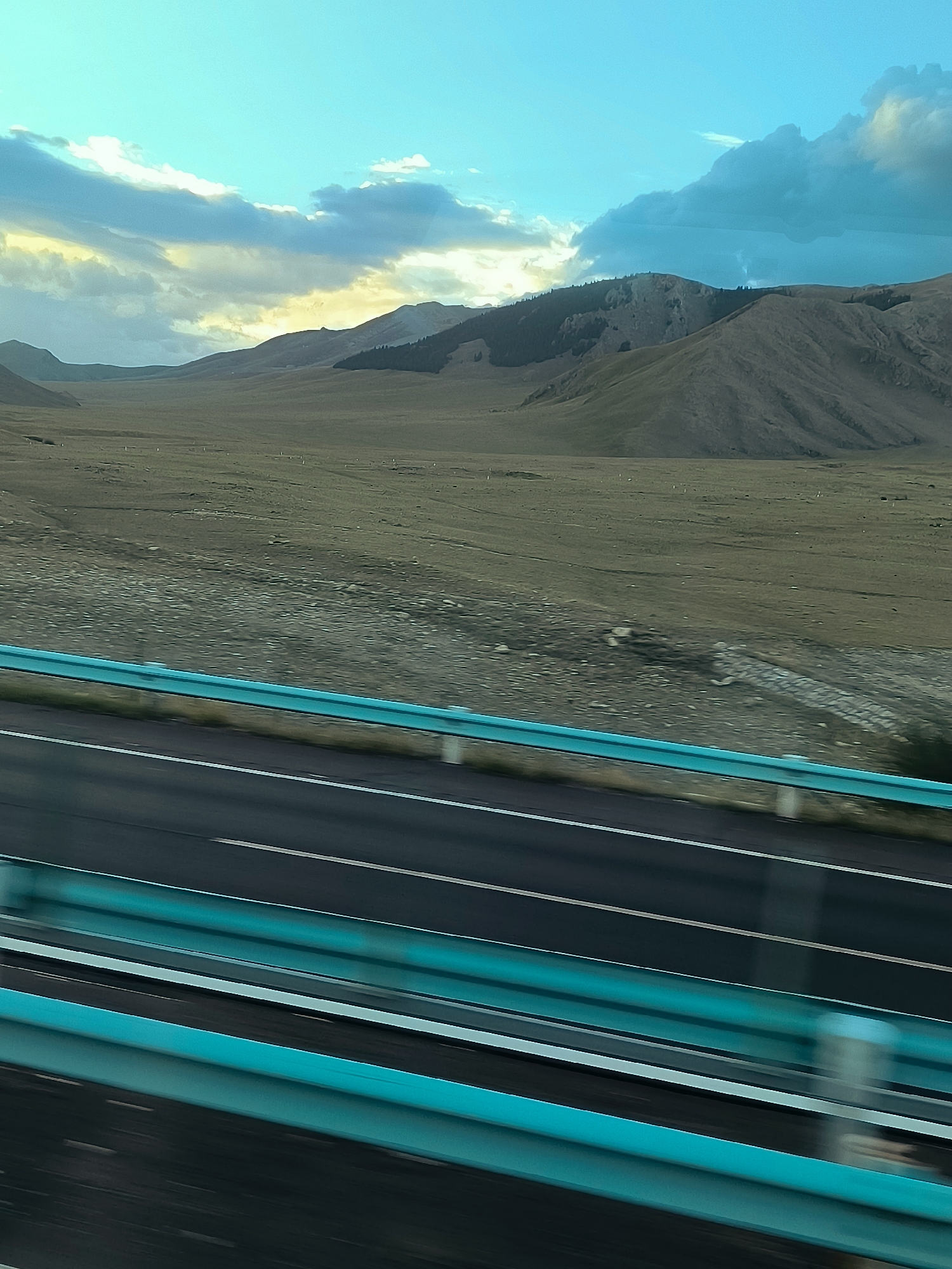 The image shows a wide, arid landscape with rolling hills under a bright, blue sky with scattered clouds. A highway stretches diagonally across the foreground, with turquoise guardrails and white lane markings blurred by motion. The vast, open terrain suggests a remote, possibly mountainous region. There are no people visible. The overall impression is one of travel through a sparsely populated natural environment.