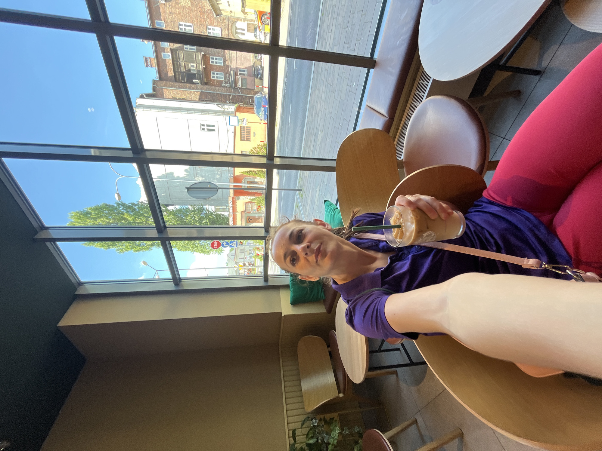A woman sits at a table inside a cafe, holding a glass of iced coffee. She is wearing a purple polo shirt and pink leggings. Through the large windows of the cafe, a street scene is visible, with buildings, cars, and trees. The woman is looking towards the camera and taking a selfie. The environment suggests a bright, sunny day.
