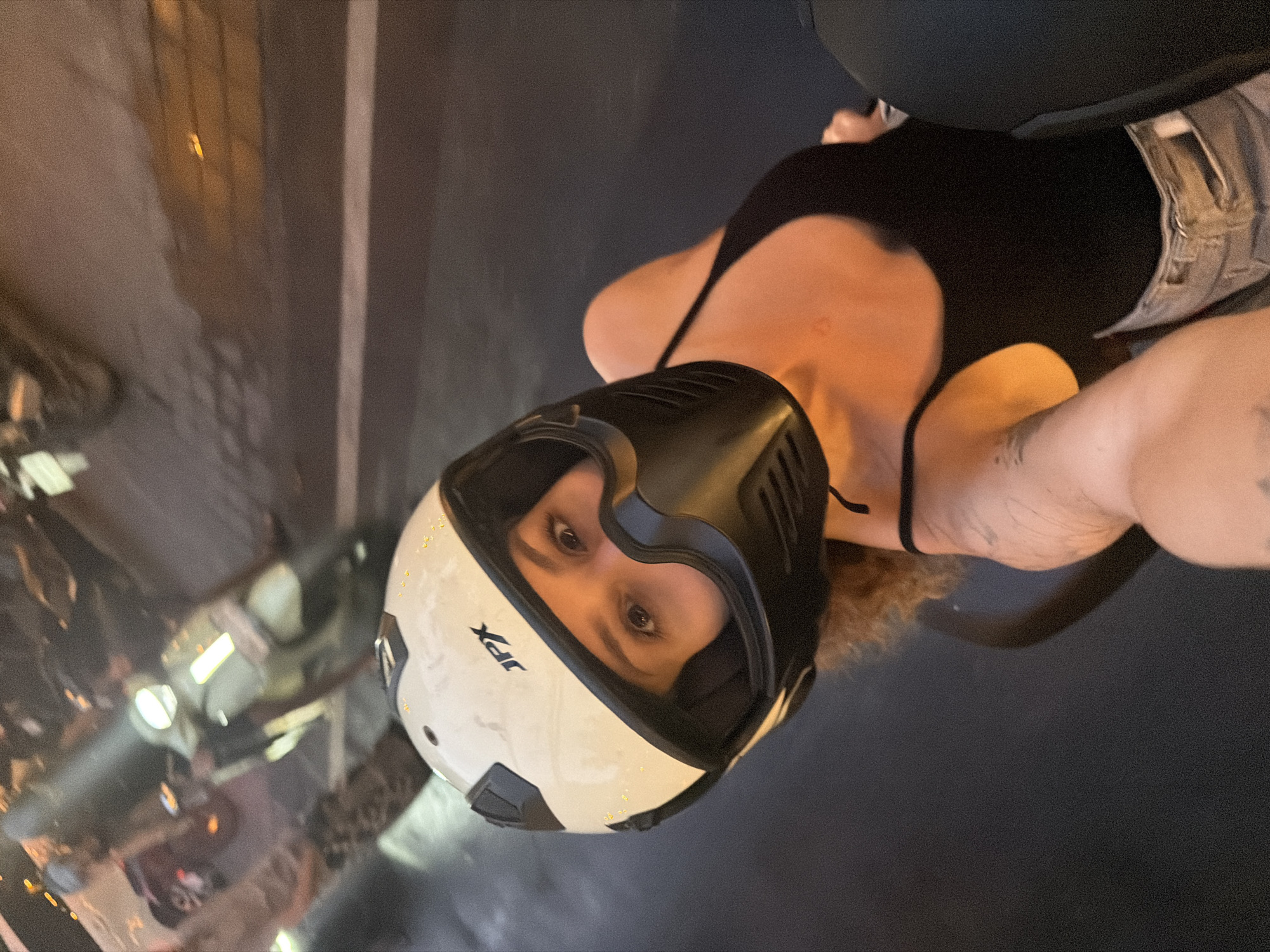 A woman wearing a white and black motorcycle helmet with a black face shield is looking at the camera while sitting on a motorcycle at night. Her eyes are visible above the face shield. She is wearing a black tank top and light-colored jeans. The background is blurred, showing street lights and other motorcycles, suggesting an urban environment. The setting is the city of Mengwi, Indonesia, at night.