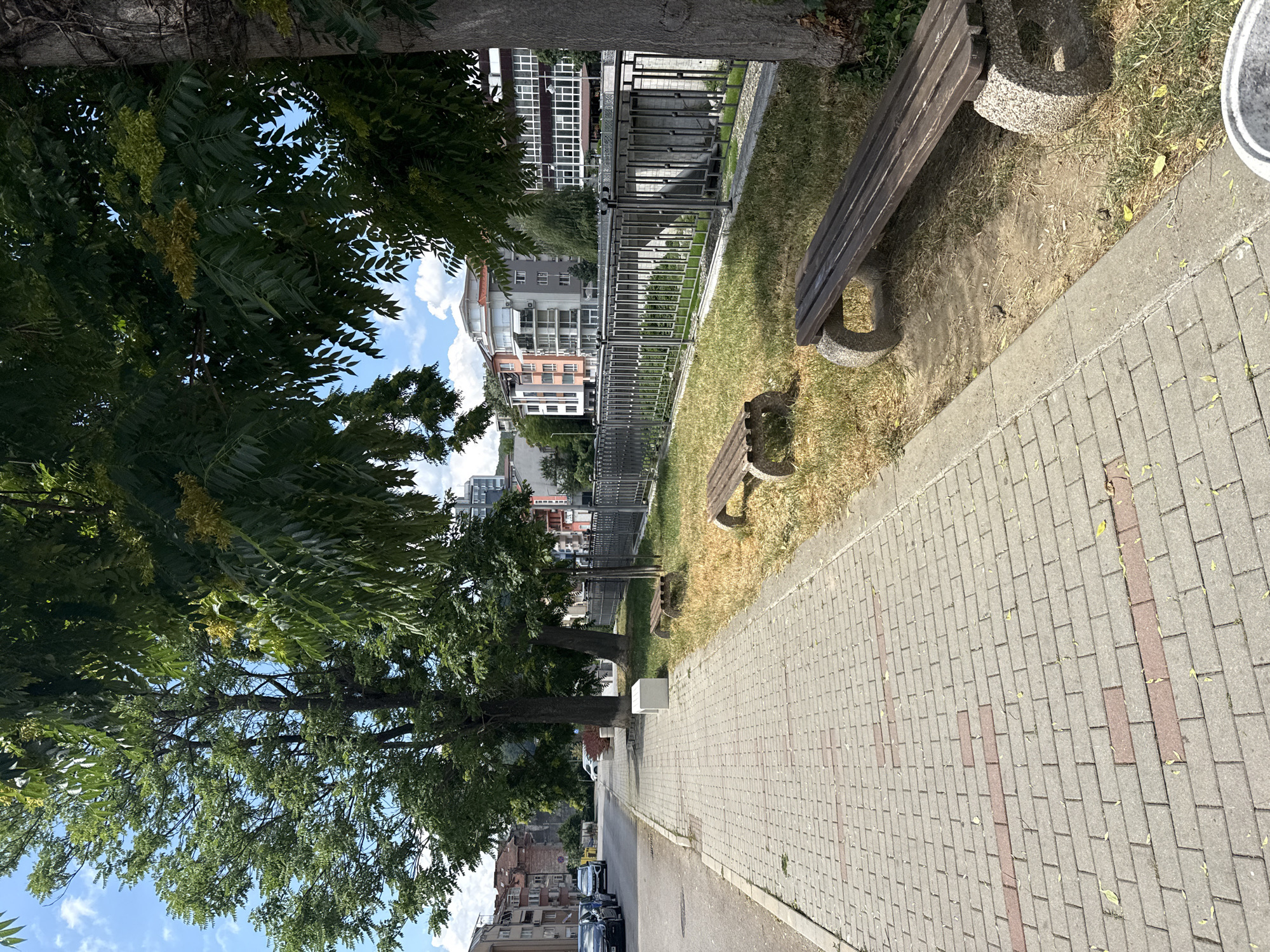 The image depicts a paved sidewalk adjacent to a grassy area with two wooden benches. The grassy area is bordered by a metal fence, which in turn overlooks a body of water. In the background, there are several multi-story buildings. The overall scene suggests an urban park or waterfront area. The specific location is identified as Gabrovo, Bulgaria, with the description likely referring to a walkway along the Yantra River. There are no people visible in the image. The day appears to be sunny with some clouds in the sky.